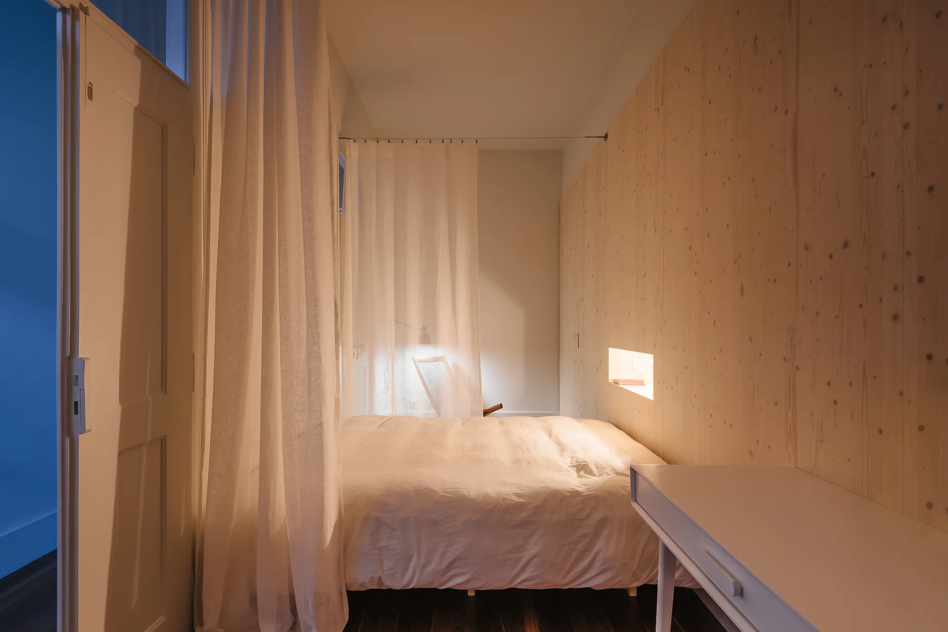 A minimalist bedroom featuring a bed with white bedding, light wood wall paneling with a lit recessed niche, and a sheer white curtain partition.