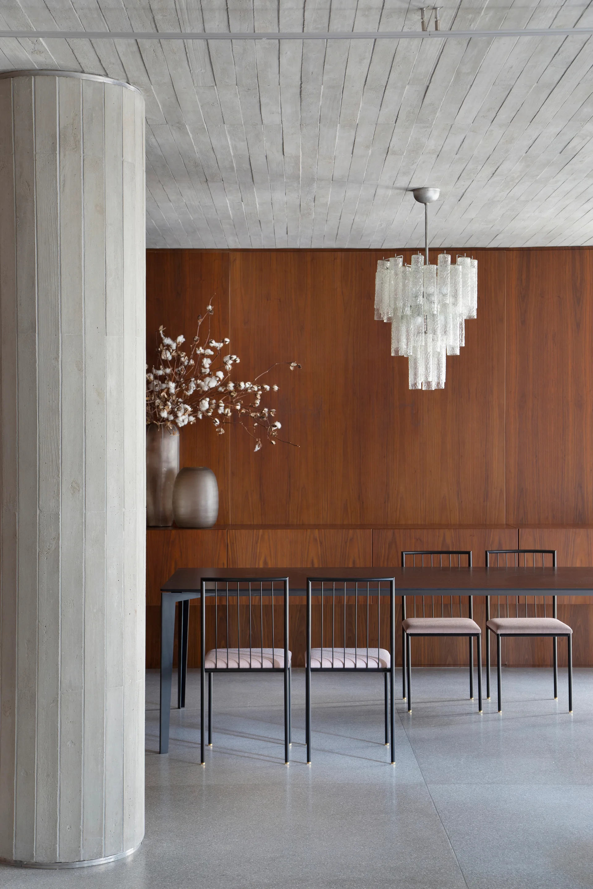 A modern dining room features black metal chairs, a tiered glass chandelier, a massive concrete column, and a warm wood-paneled wall.