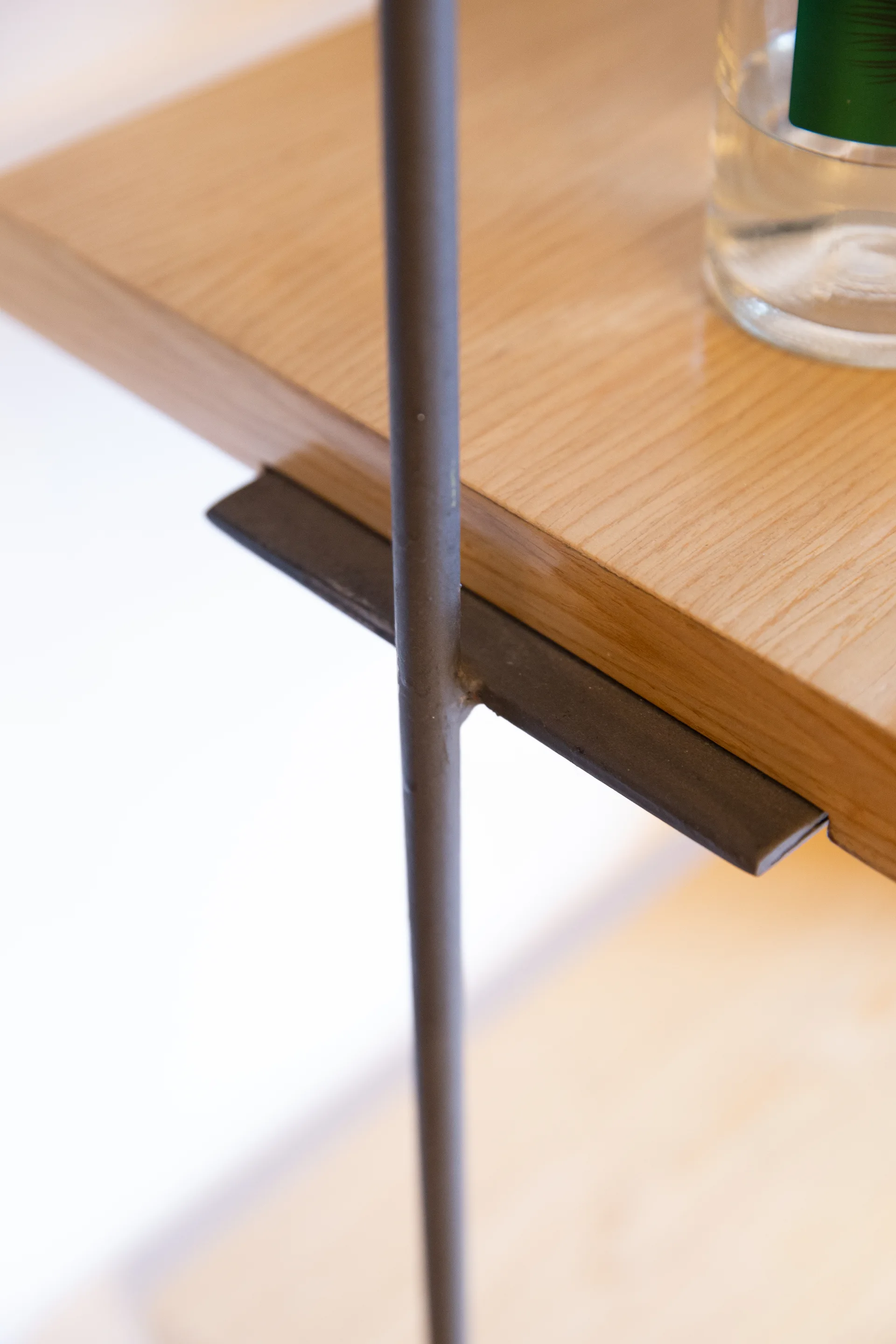 A close-up shot of a light wood shelf resting on a dark matte metal support bracket welded to a vertical steel rod.