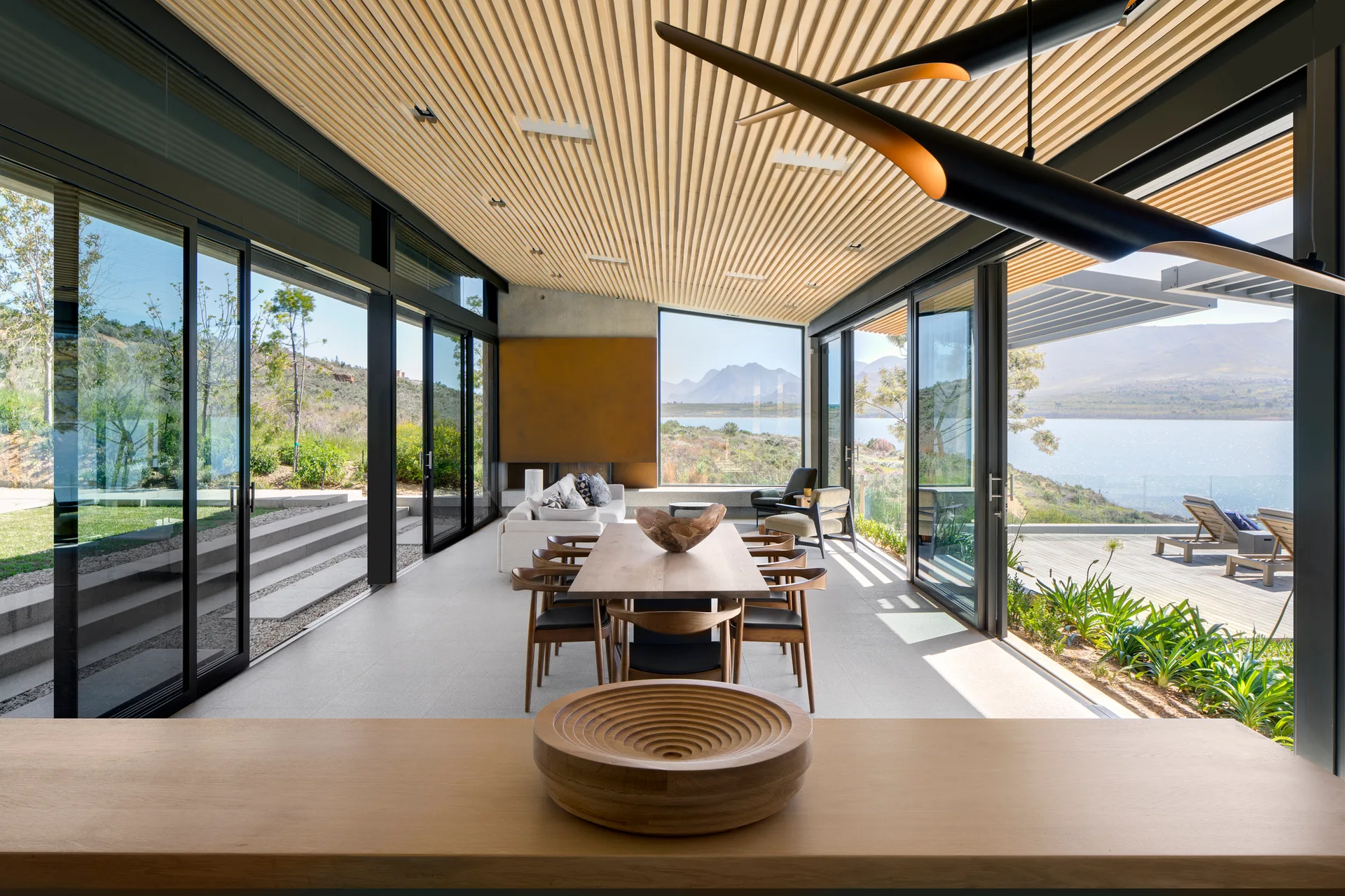 A minimalist dining room features a long wooden table, slatted wood ceiling, and floor-to-ceiling glass doors opening to a scenic lake view.