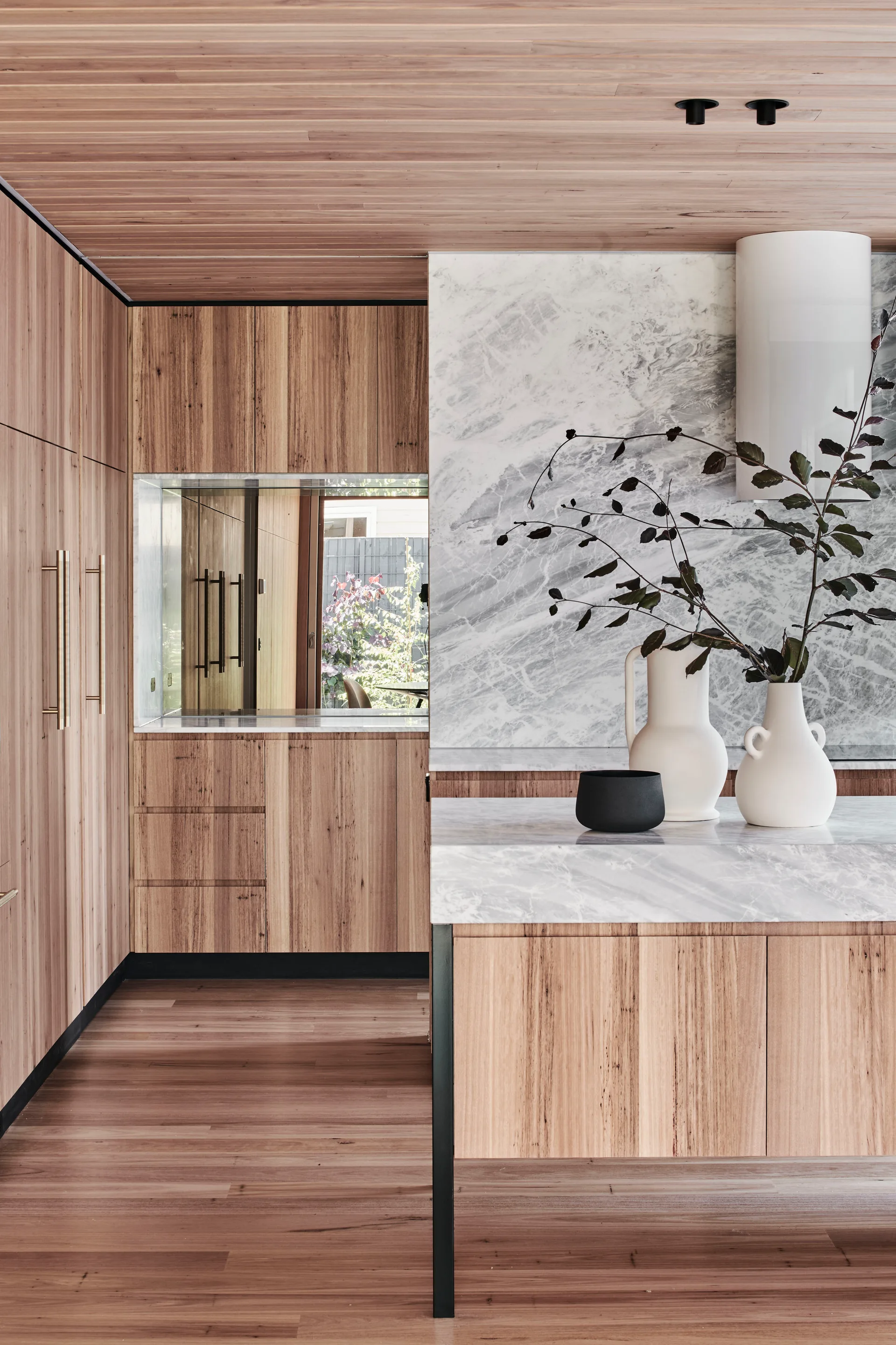 Modern kitchen with warm wood cabinetry, a matching slatted ceiling, a heavily veined marble backsplash, and white ceramic vases on the island.