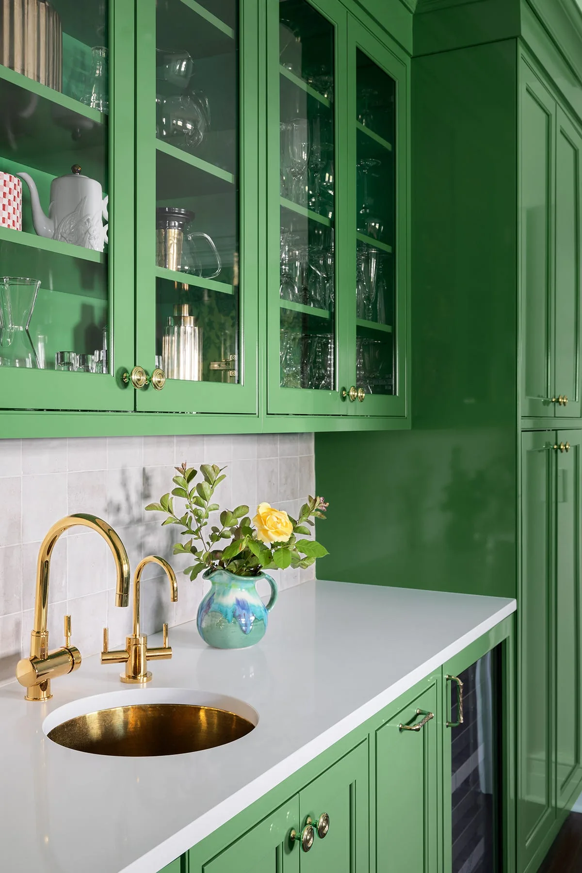 A close-up of a green wet bar featuring gloss-painted cabinetry, a white countertop, an undermount gold sink, and a brass faucet.