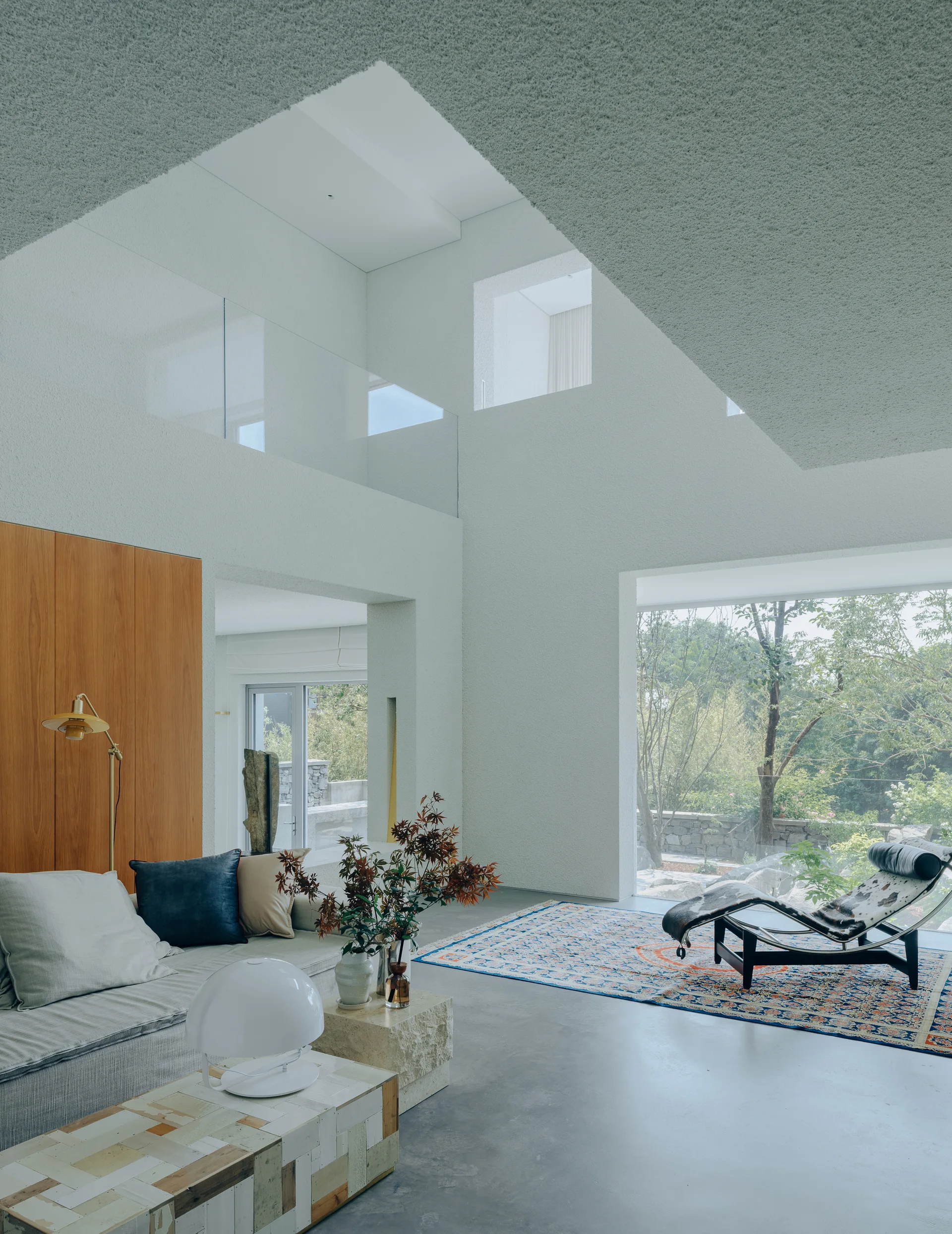A double-height modern living room features textured white walls, a concrete floor, a low sofa, a patterned rug, and a large floor-to-ceiling window.