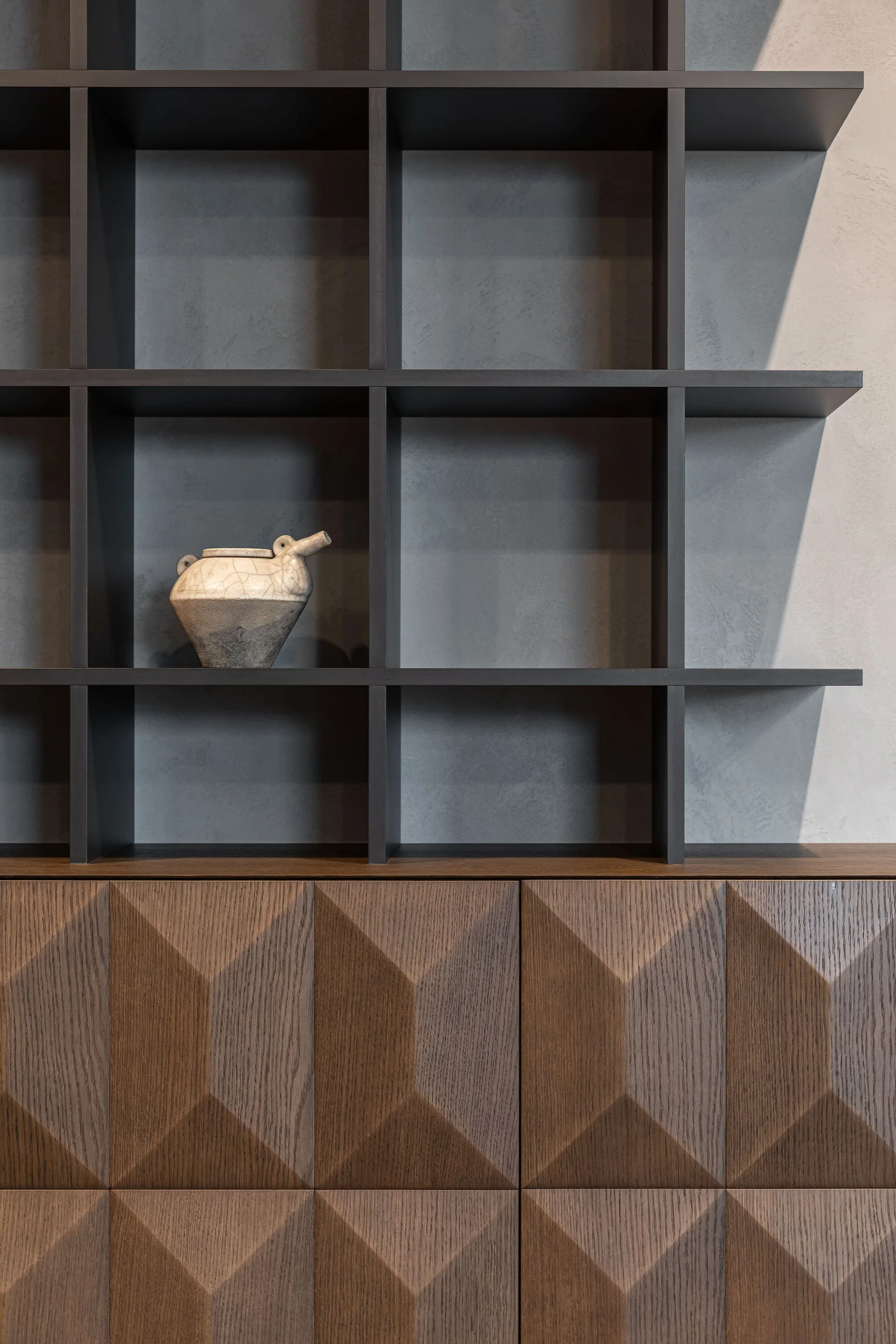 Dark grey open grid shelves holding a single ceramic vessel above a dark wood cabinet with dimensional geometric-patterned doors.