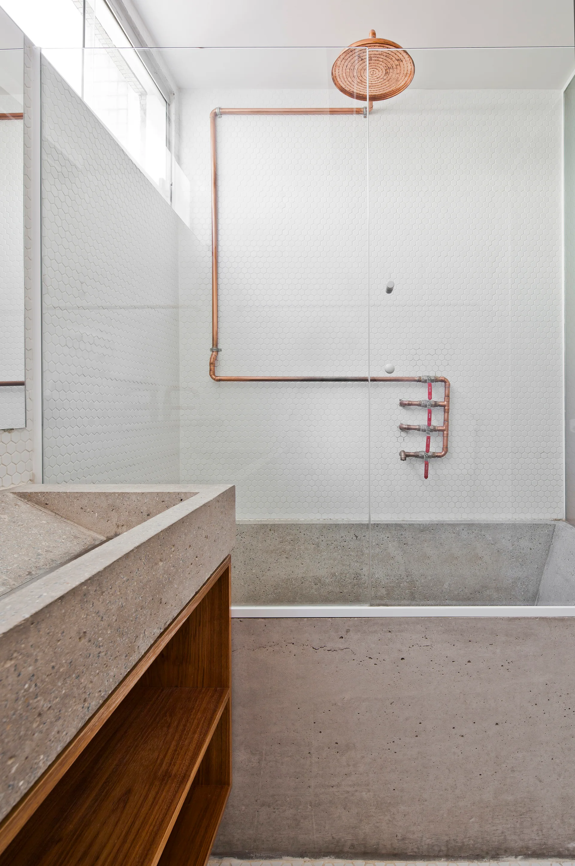 A minimalist bathroom features a textured concrete sink on a wood vanity beside a glass shower with exposed copper plumbing against white hexagon tiles.