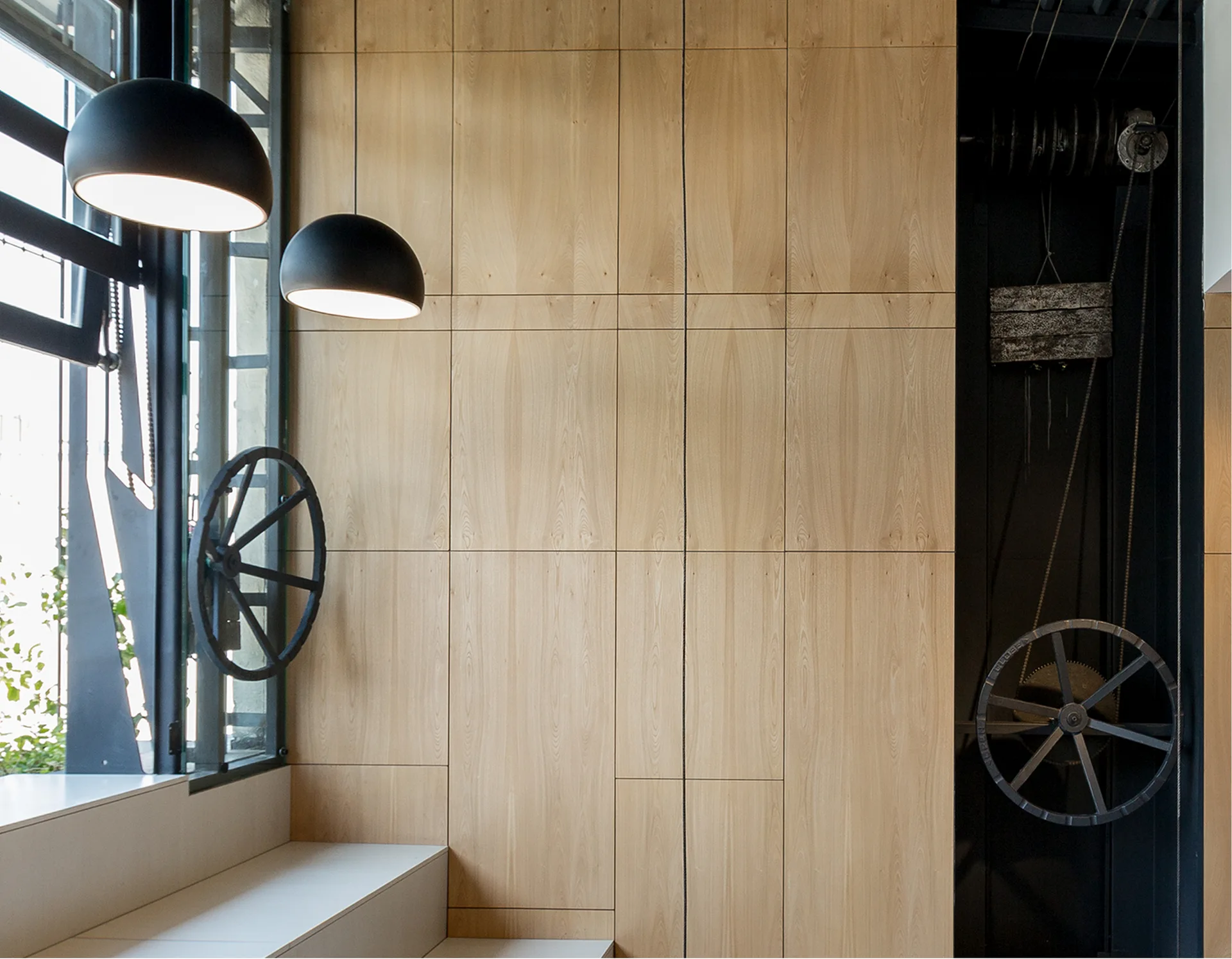 Two black dome pendant lights hang beside a light wood paneled wall and an exposed black mechanical pulley system.