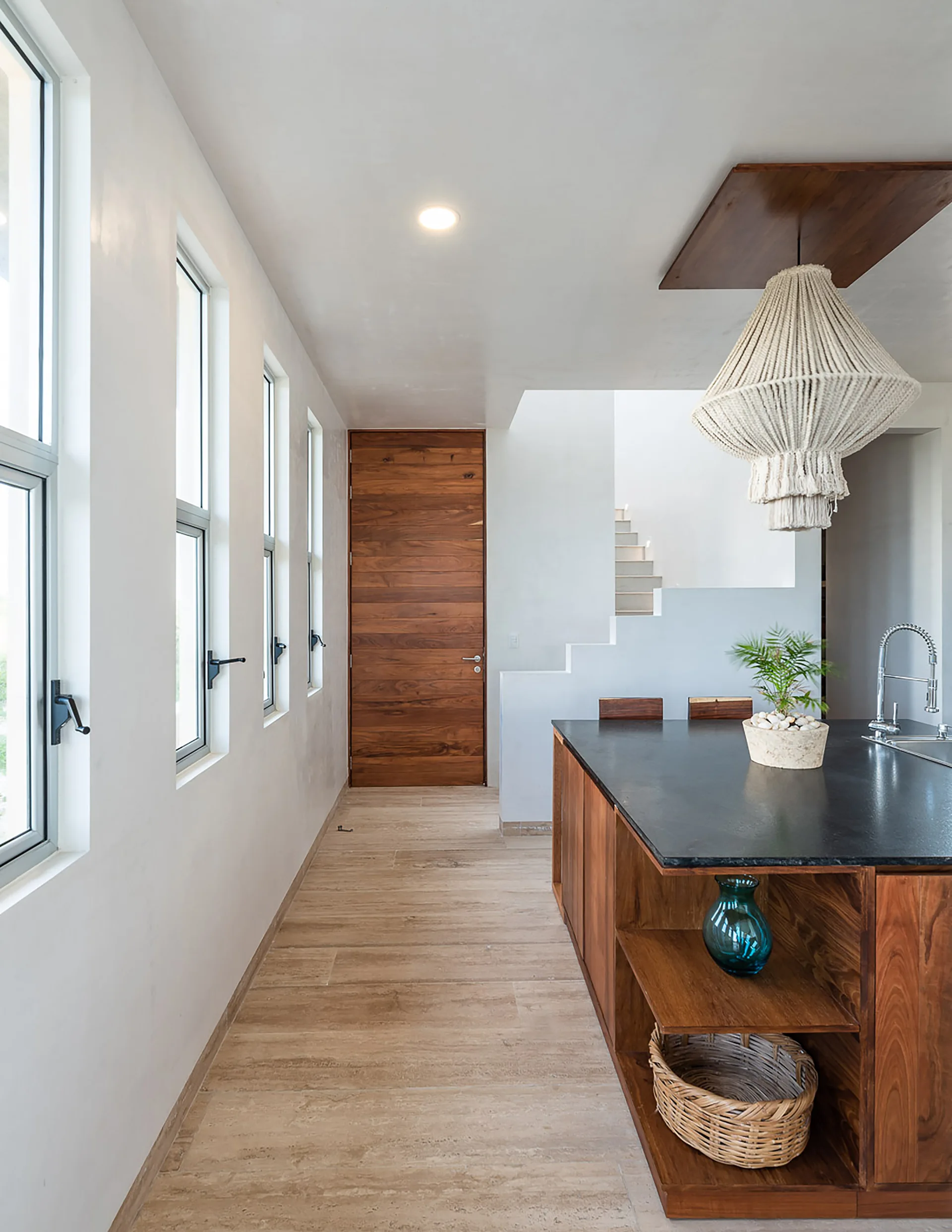 A modern kitchen featuring a wood island with a black countertop, a large macrame pendant light, and a row of narrow windows on a white wall.