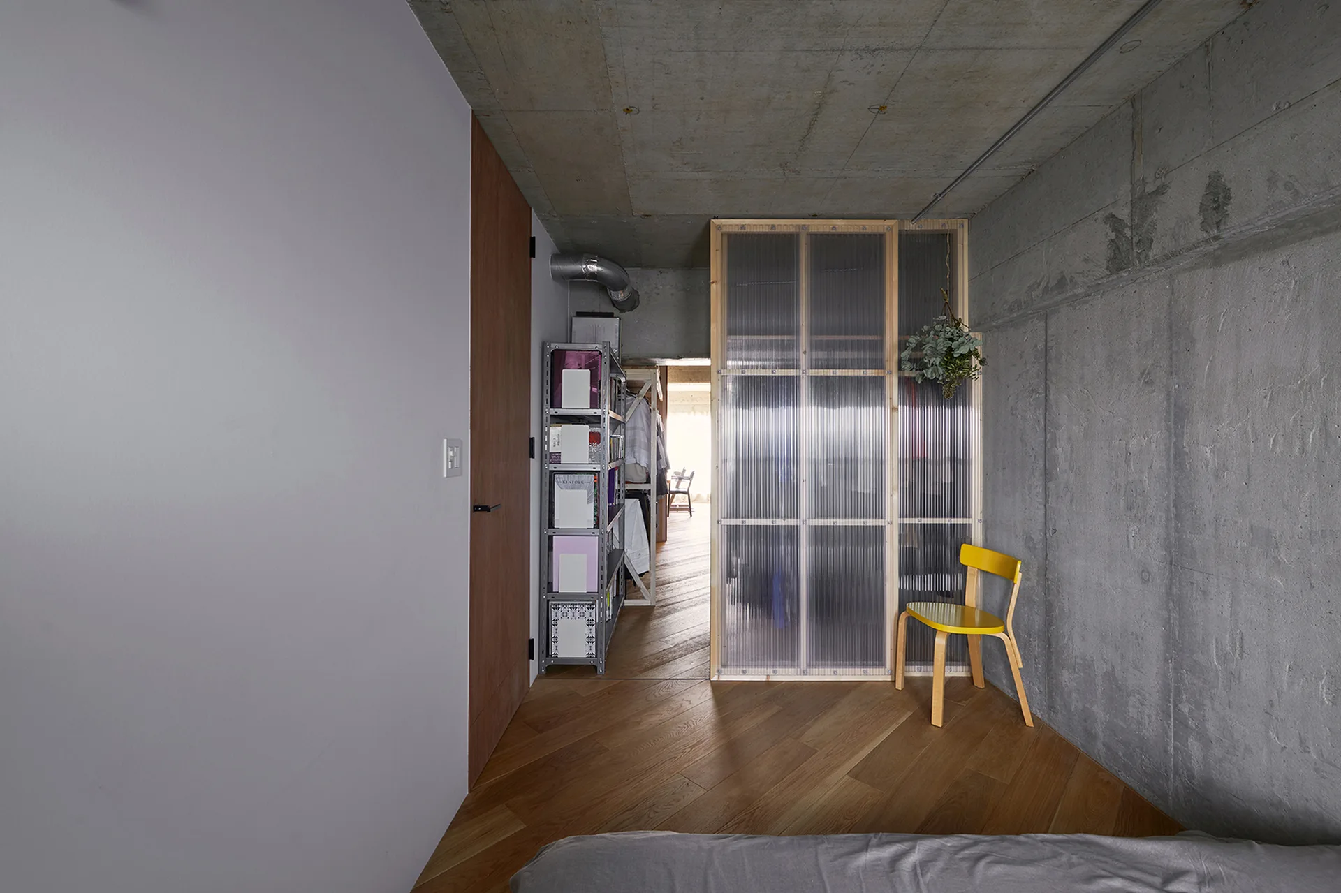 An industrial bedroom featuring exposed concrete walls, herringbone wood flooring, a yellow chair, and a translucent corrugated room divider.