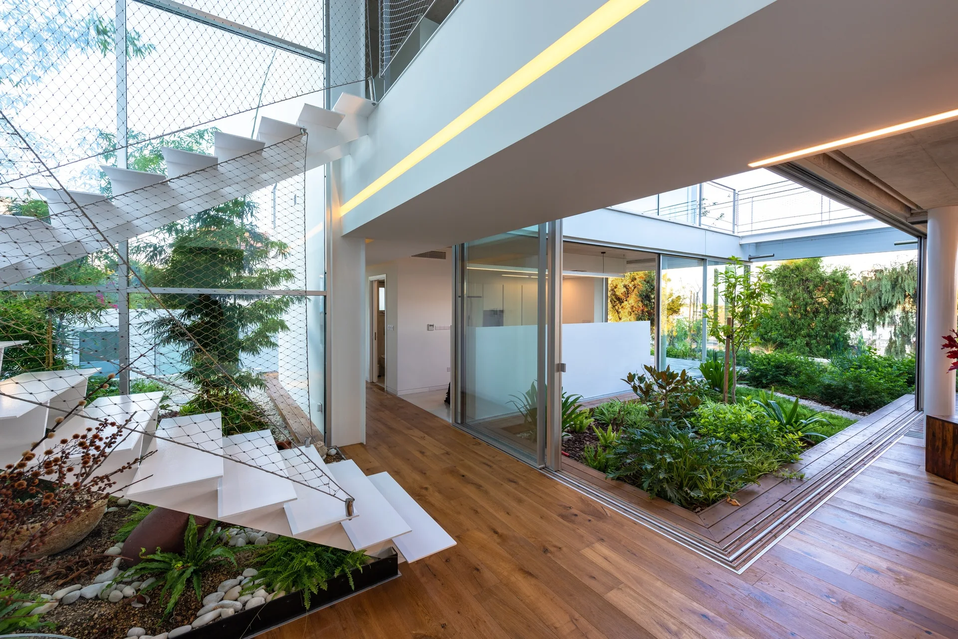 A white floating staircase with wire mesh railing rises next to an indoor garden courtyard and warm wood floors in a modern home.
