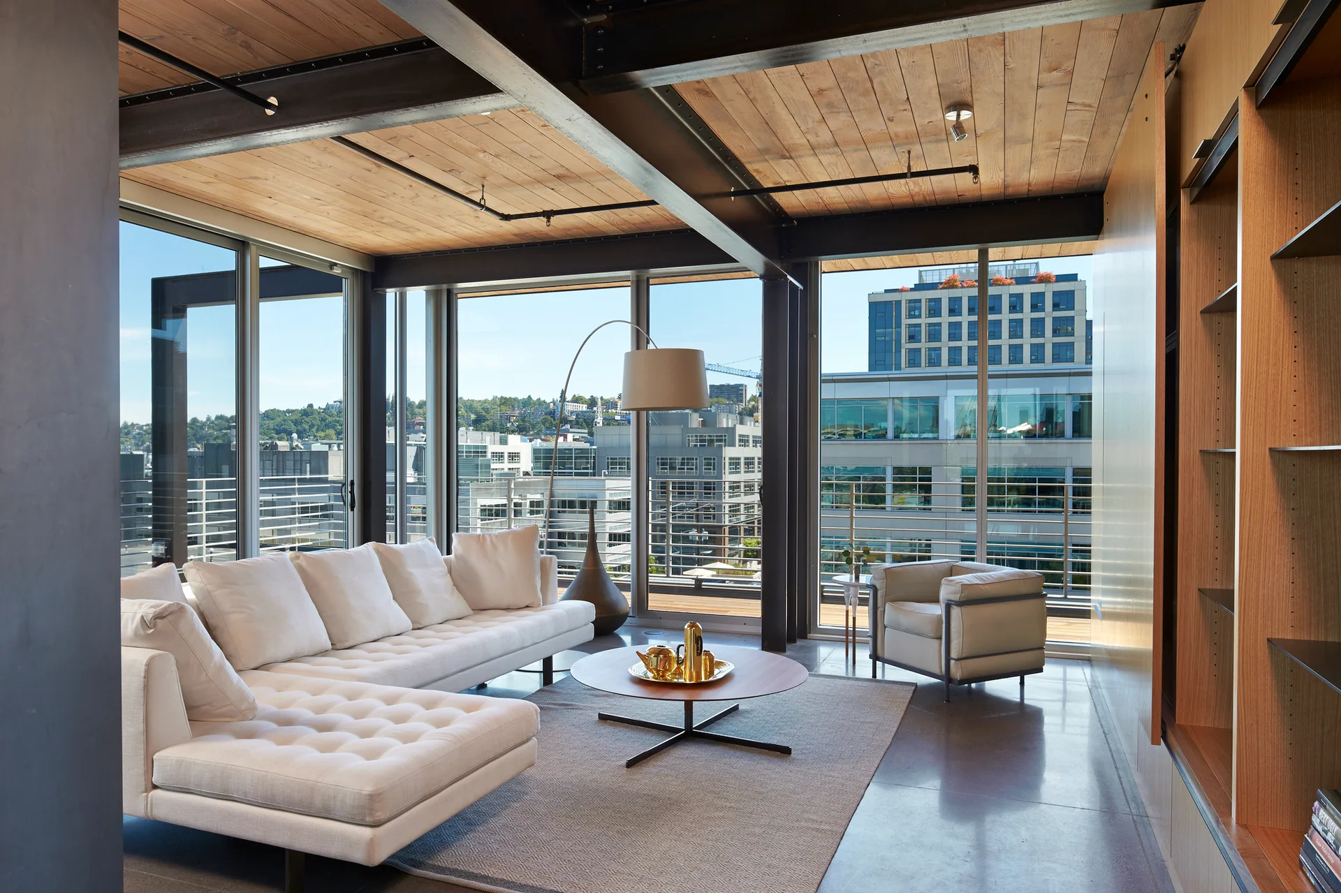 Modern living room with a white tufted sectional sofa, round wooden coffee table, exposed steel beams, wood ceiling, and floor-to-ceiling windows.