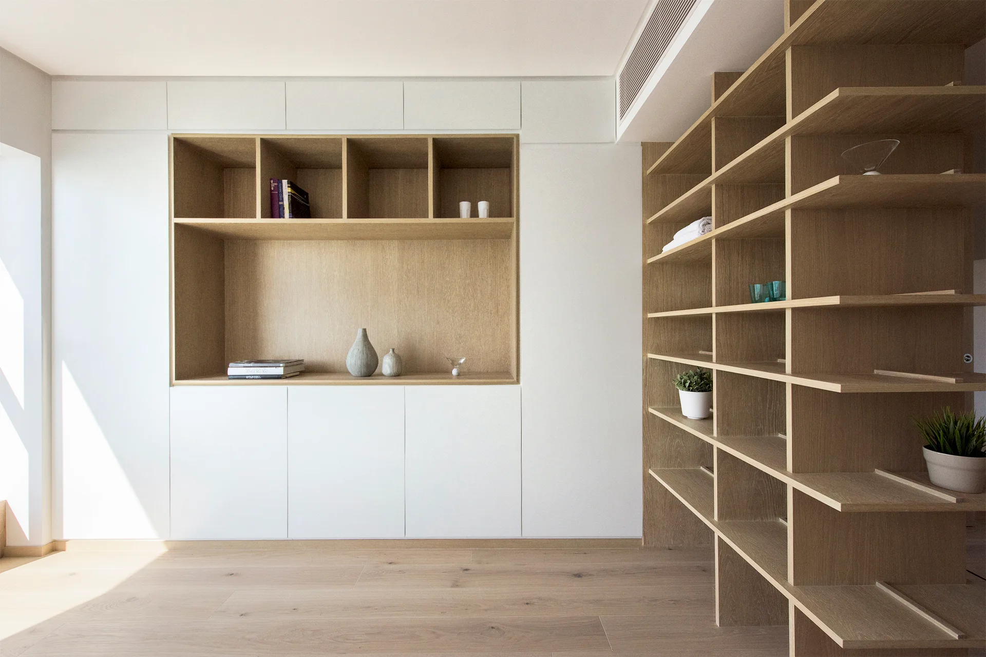 Minimalist white built-in cabinets feature a recessed light wood shelving niche next to a matching tall wooden bookshelf on light oak floors.
