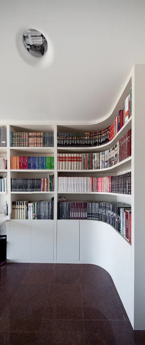 A white custom built-in bookshelf curves around a corner in a room with dark tiled floors and books neatly organized on open shelves.