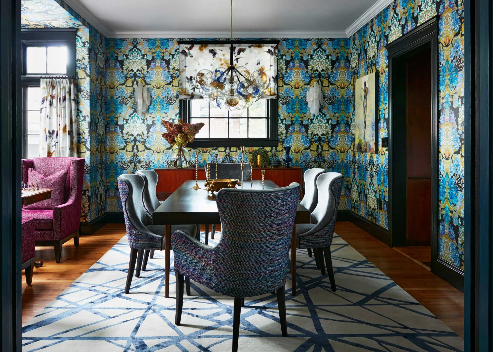 A maximalist dining room features bold blue and yellow patterned wallpaper, a dark wood table with textured upholstered chairs, and a glass globe chandelier.