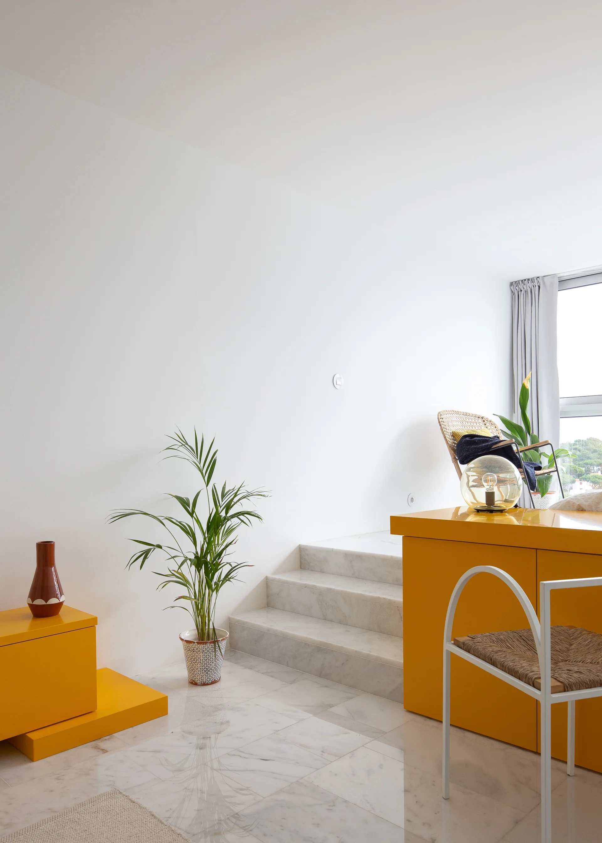 A minimalist white living area features glossy yellow cabinets, a white metal-framed chair, a potted palm, and a light grey marble staircase.