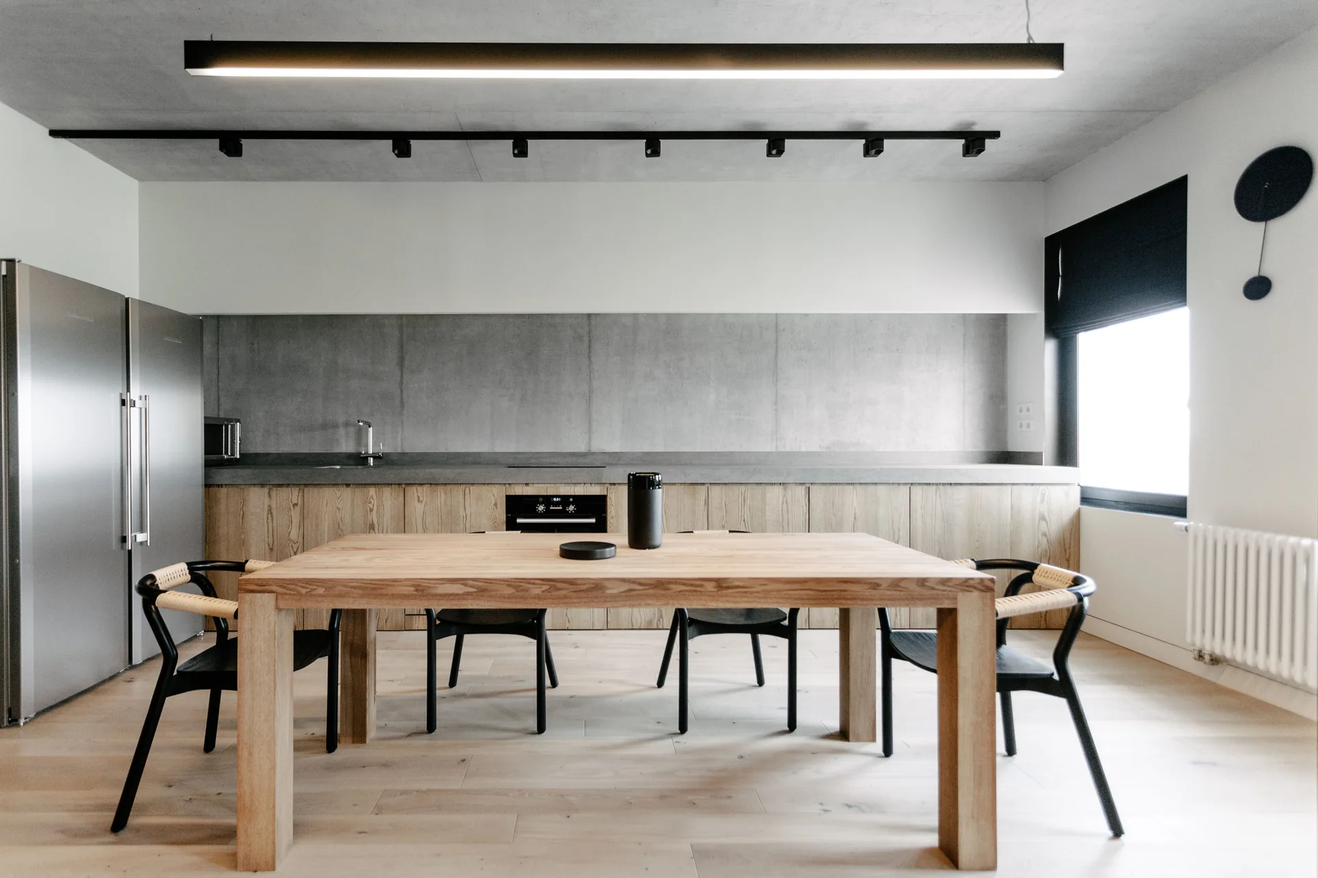 A minimalist kitchen features light wood cabinets, a concrete backsplash, and a natural wood dining table with black chairs.