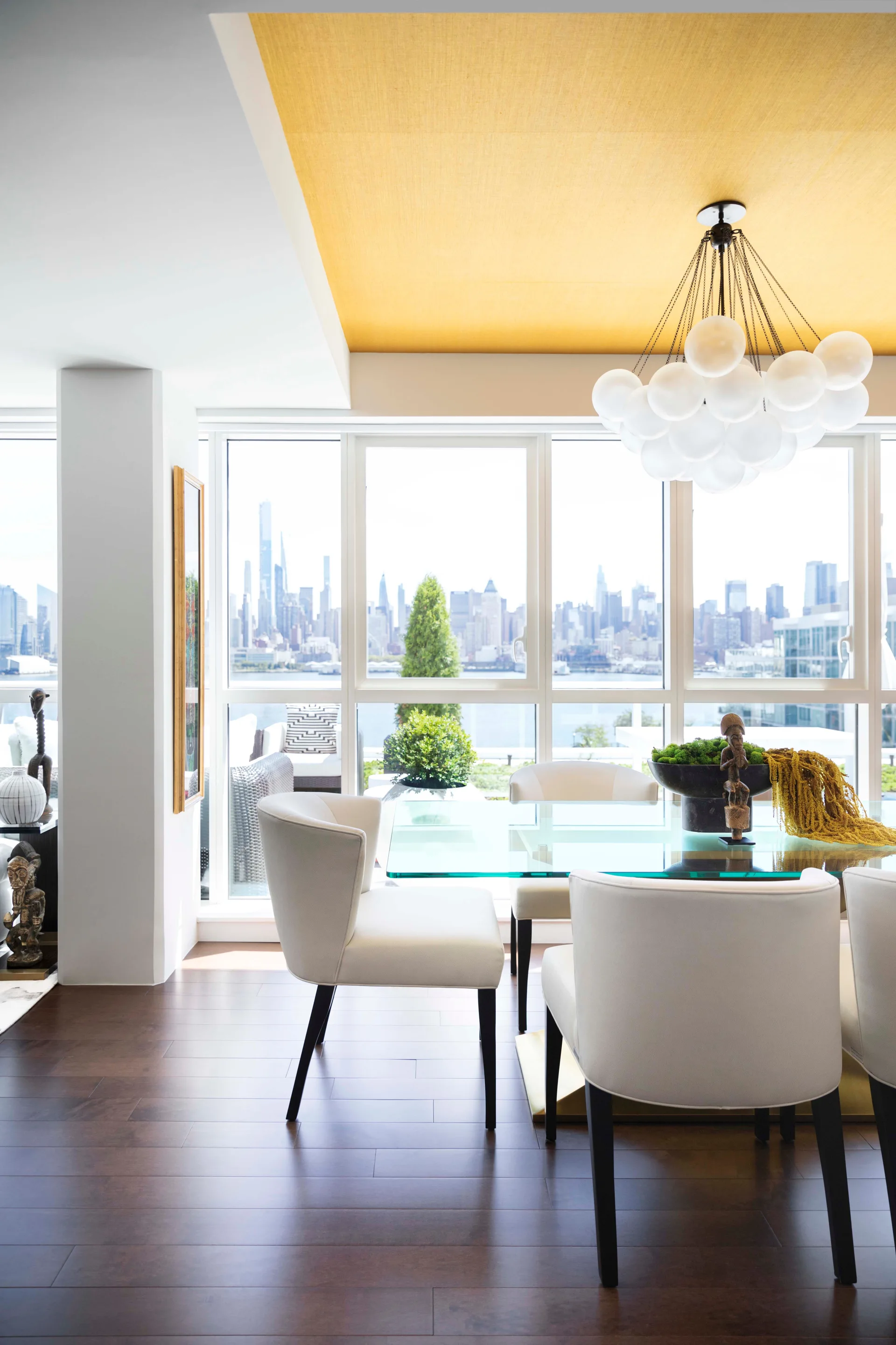 A modern dining room featuring white upholstered chairs around a glass dining table, a bubble chandelier, and a yellow textured ceiling panel.