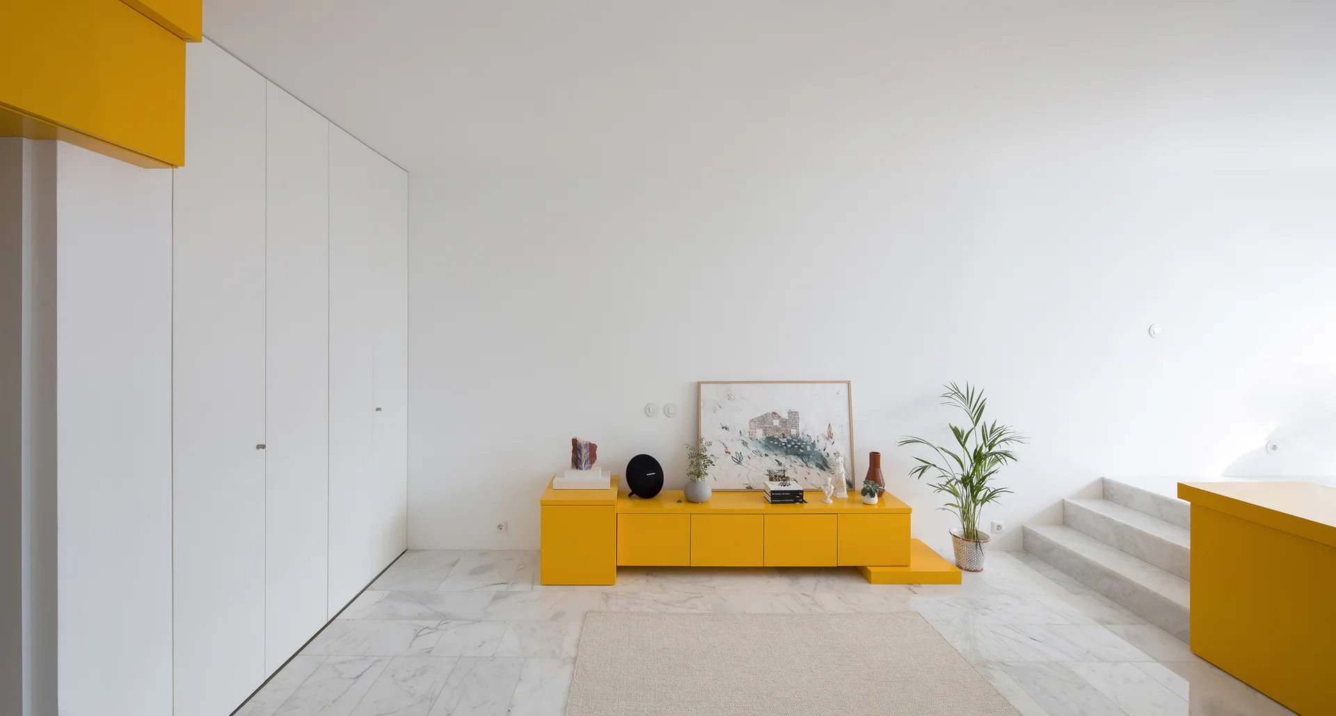 A minimalist living room featuring a low-profile bright yellow media console, tall seamless white storage cabinets, and polished white marble flooring.