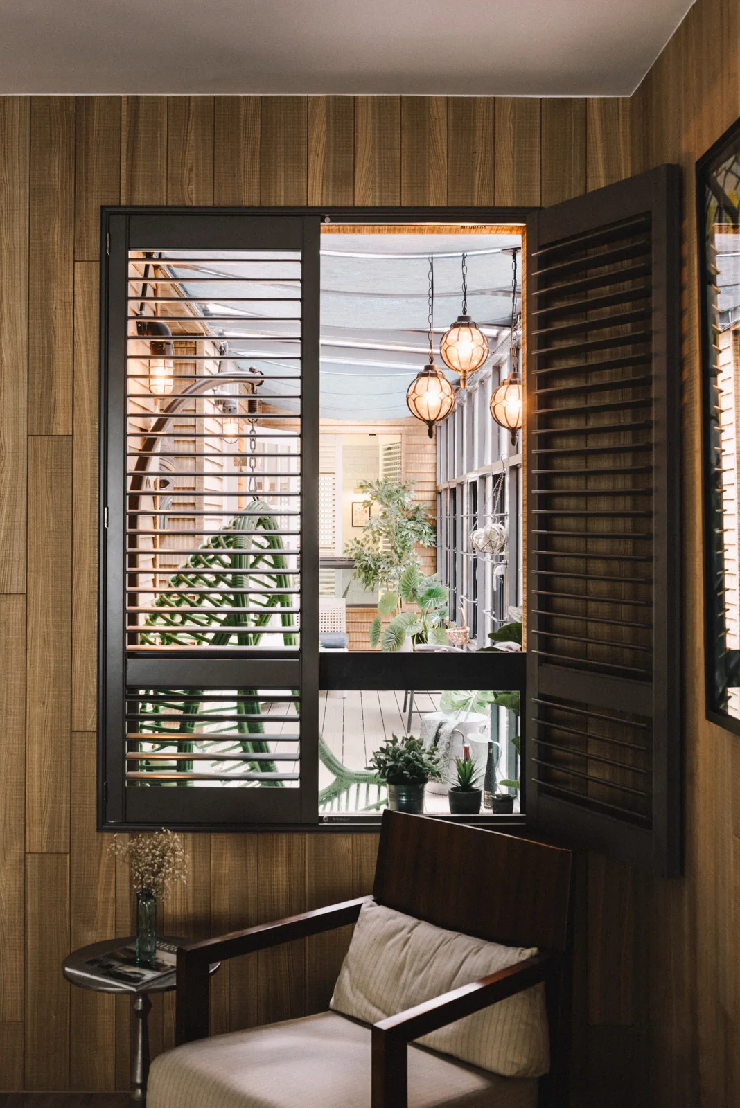 A cozy interior corner features vertical wood plank paneling, a cushioned armchair, and an open shuttered window looking out to a covered patio.