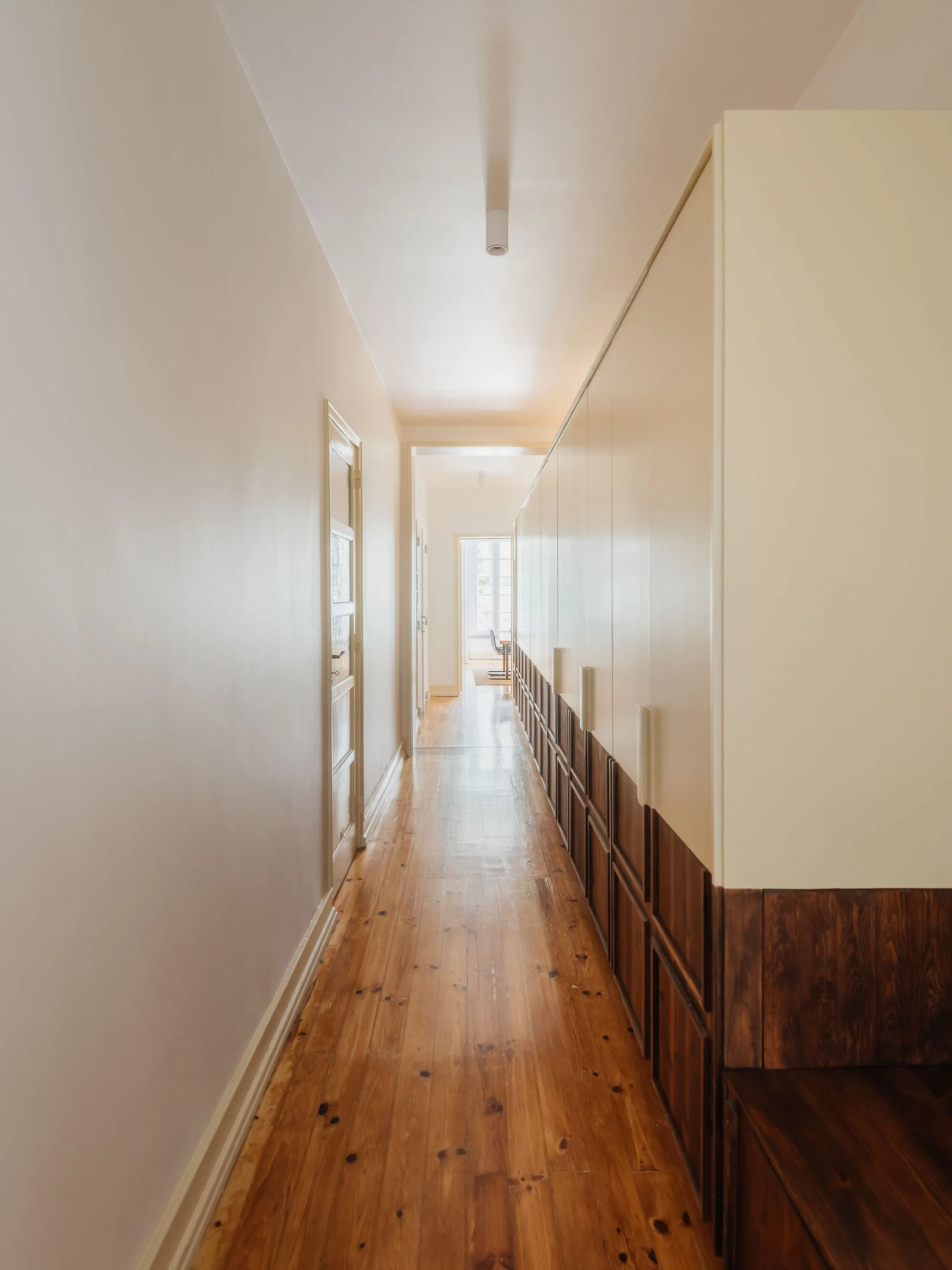 A long residential hallway featuring wide-plank wood flooring and a wall of built-in cabinets with white upper doors and dark wood lower panels.