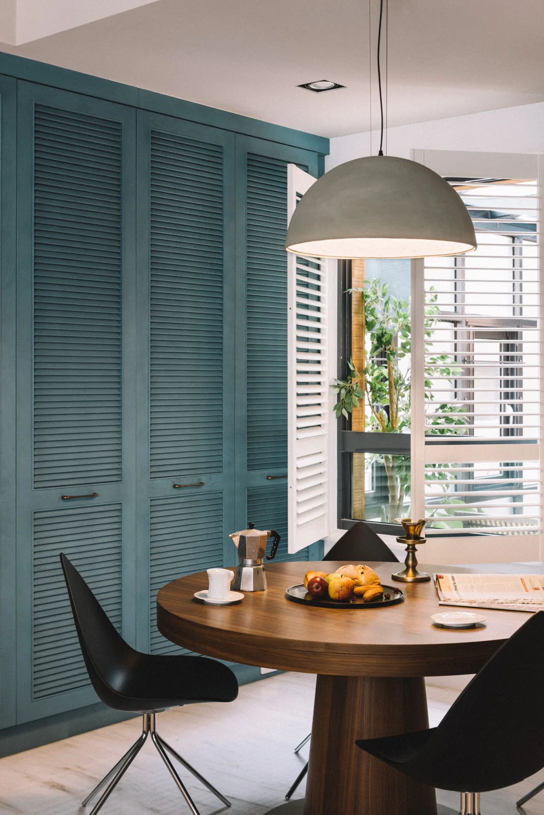 A round wood dining table with black modern chairs sits before a large wall of teal louvered cabinetry under a gray dome pendant light.