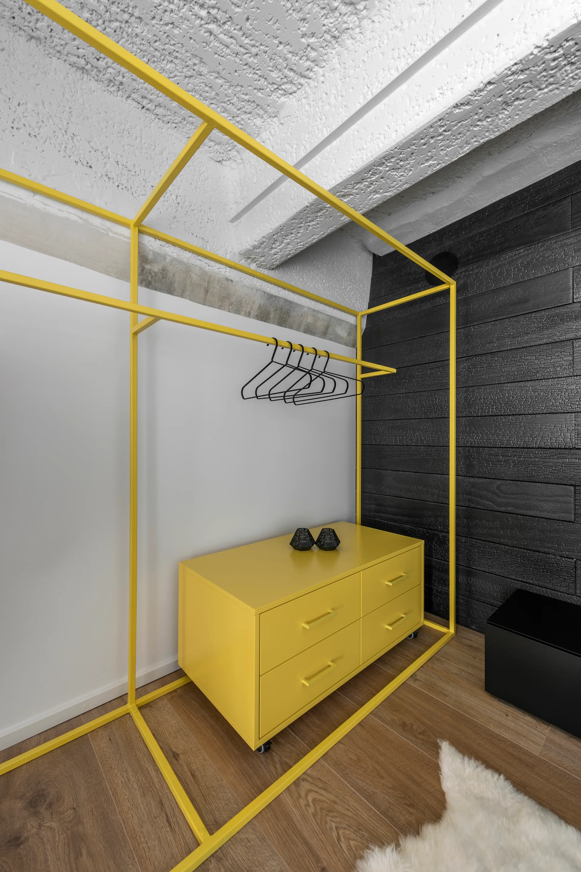 A bright yellow metal wardrobe frame and matching dresser stand against a textured black charred wood plank wall on hardwood floors.