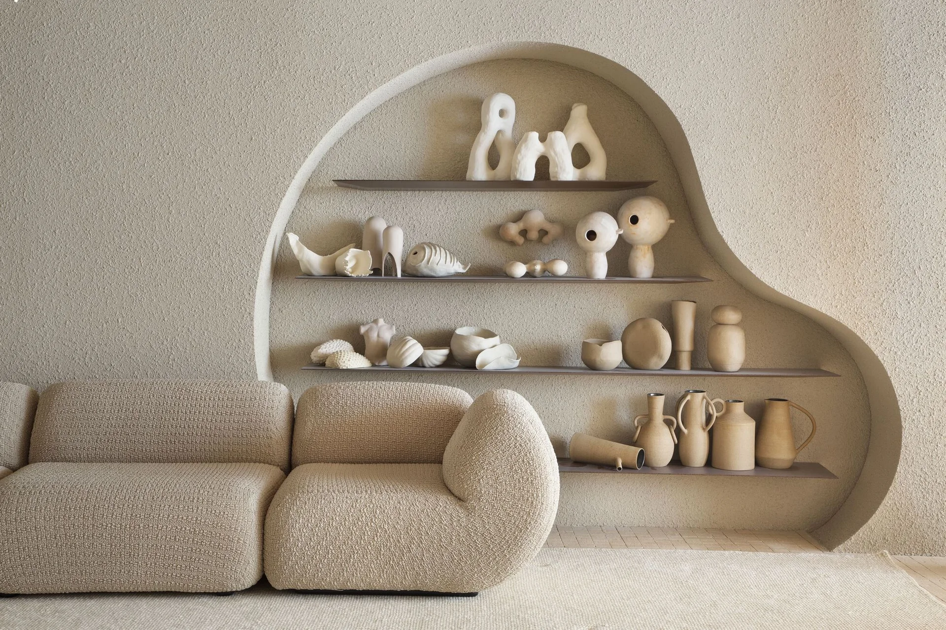 A curved beige boucle sofa sits before a heavily textured plaster wall featuring an asymmetrical arched niche with shelves holding ceramic art.