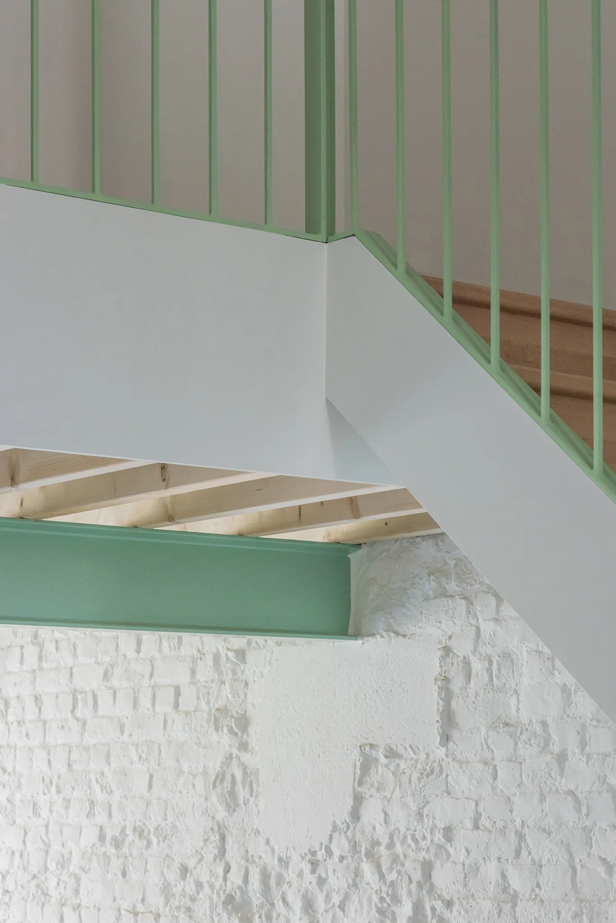 A structural staircase features a mint green metal handrail and steel beam set against a textured white painted brick wall and exposed timber joists.