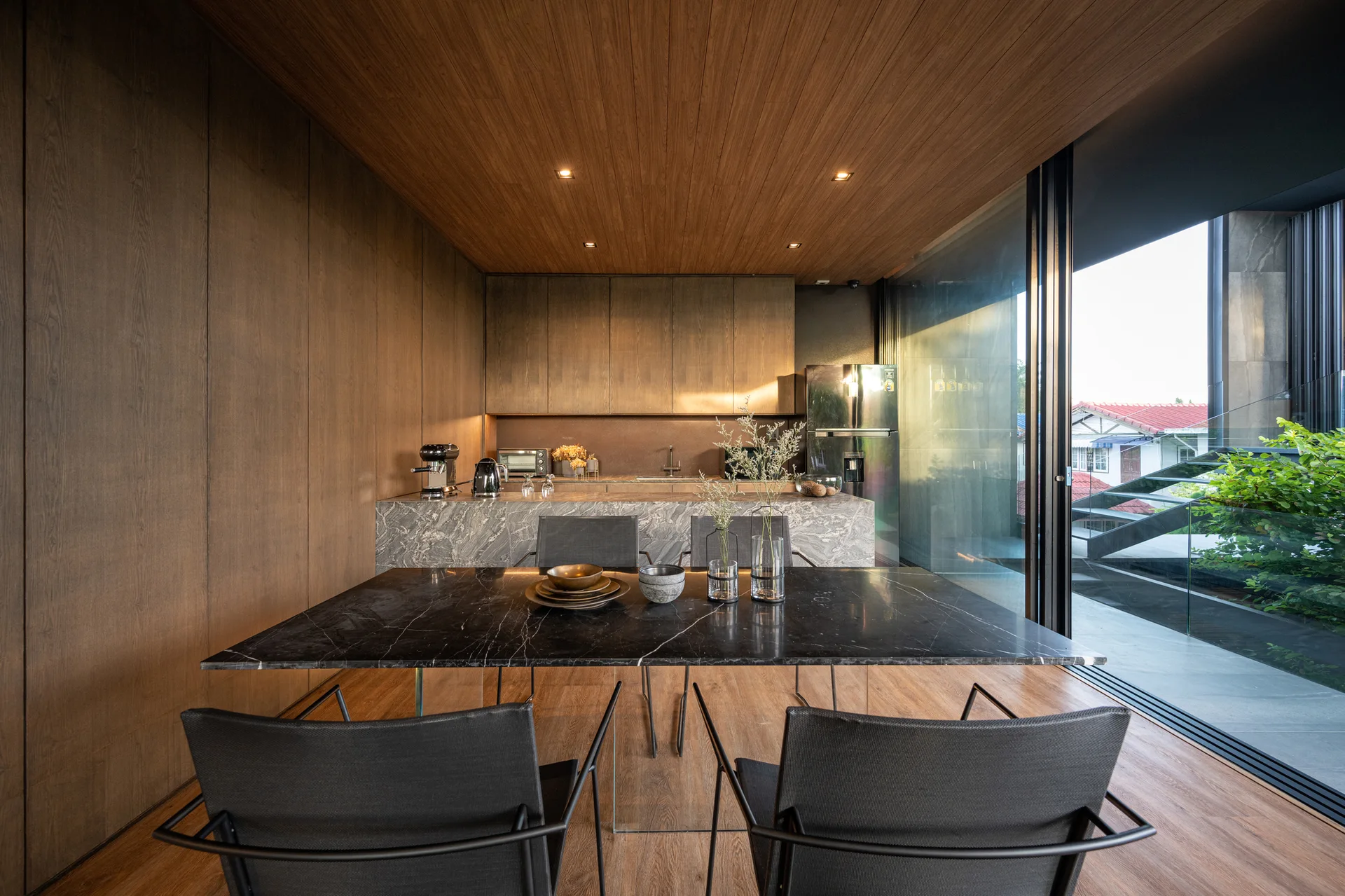A modern dining area features a black marble table and chairs set against wood-paneled walls, a matching wood ceiling, and a grey marble kitchen island.