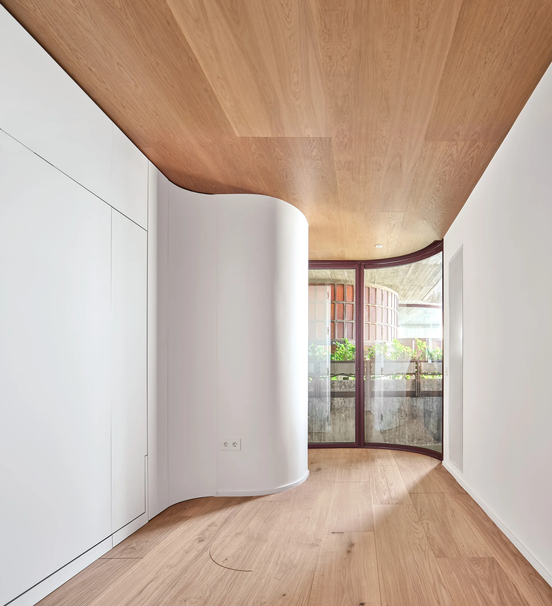 A minimalist hallway features light wood floors and ceiling panels, a curved white partition wall with built-in storage, and a curved glass balcony door.
