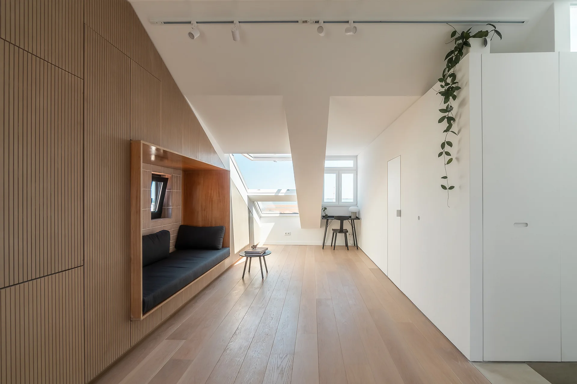 A minimalist attic room with light wood floors, a sloped ceiling with skylight, and a slatted wood wall with a recessed black bench.