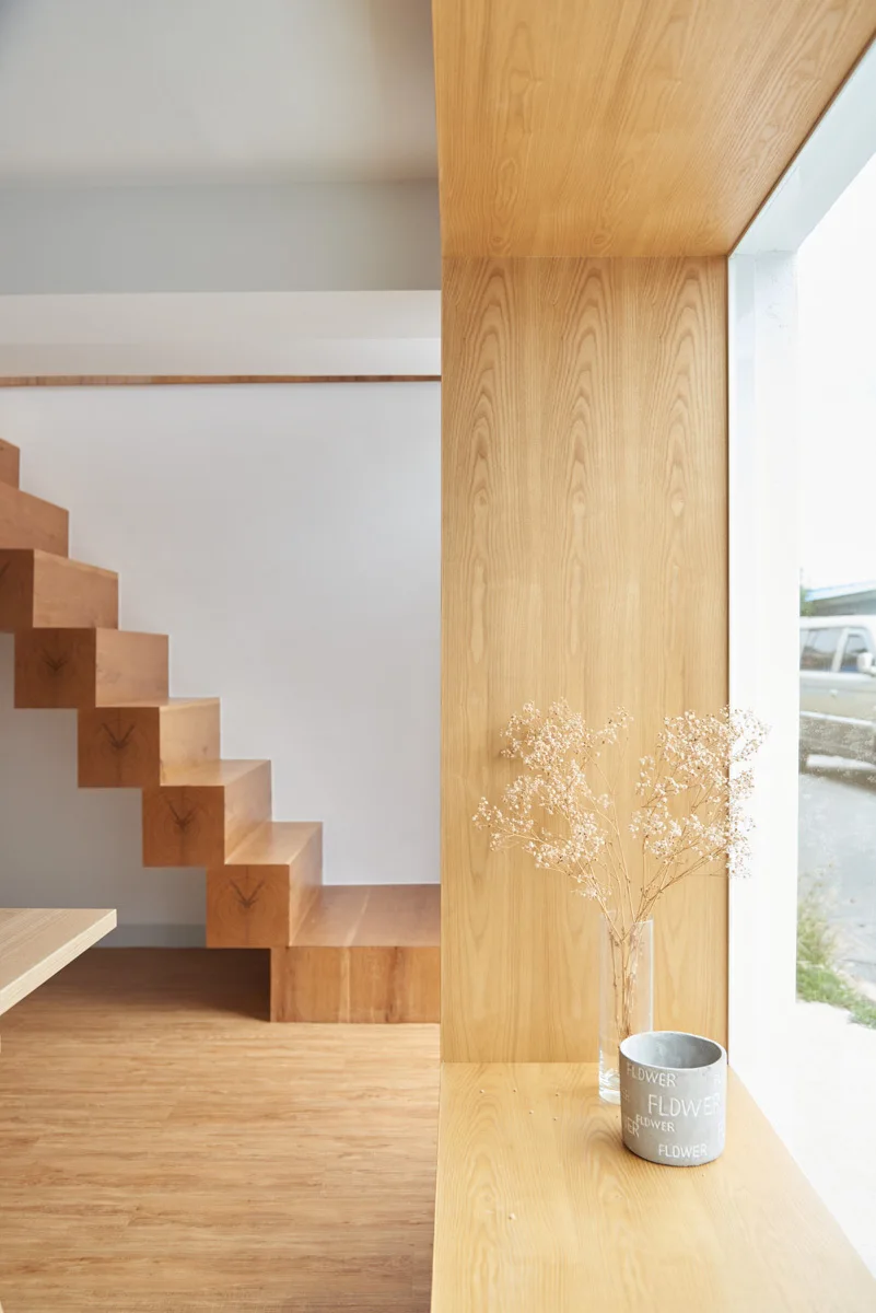 A minimalist interior featuring a floating light wood staircase and an adjacent wood-paneled window nook holding a glass vase and small pot.