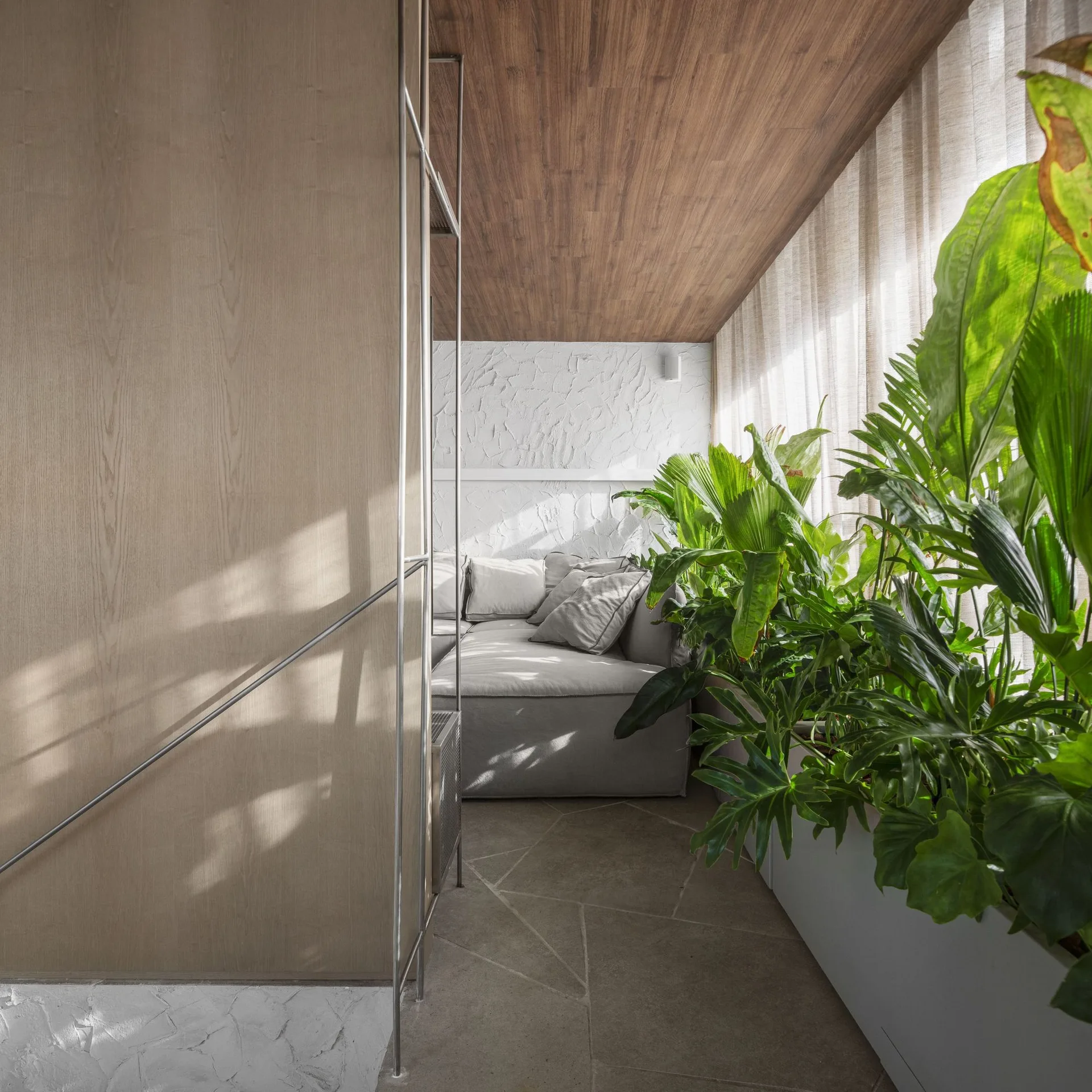 A modern seating nook features a grey upholstered sofa in a corner with a wood-planked ceiling, textured white wall, and a long white planter of green plants.