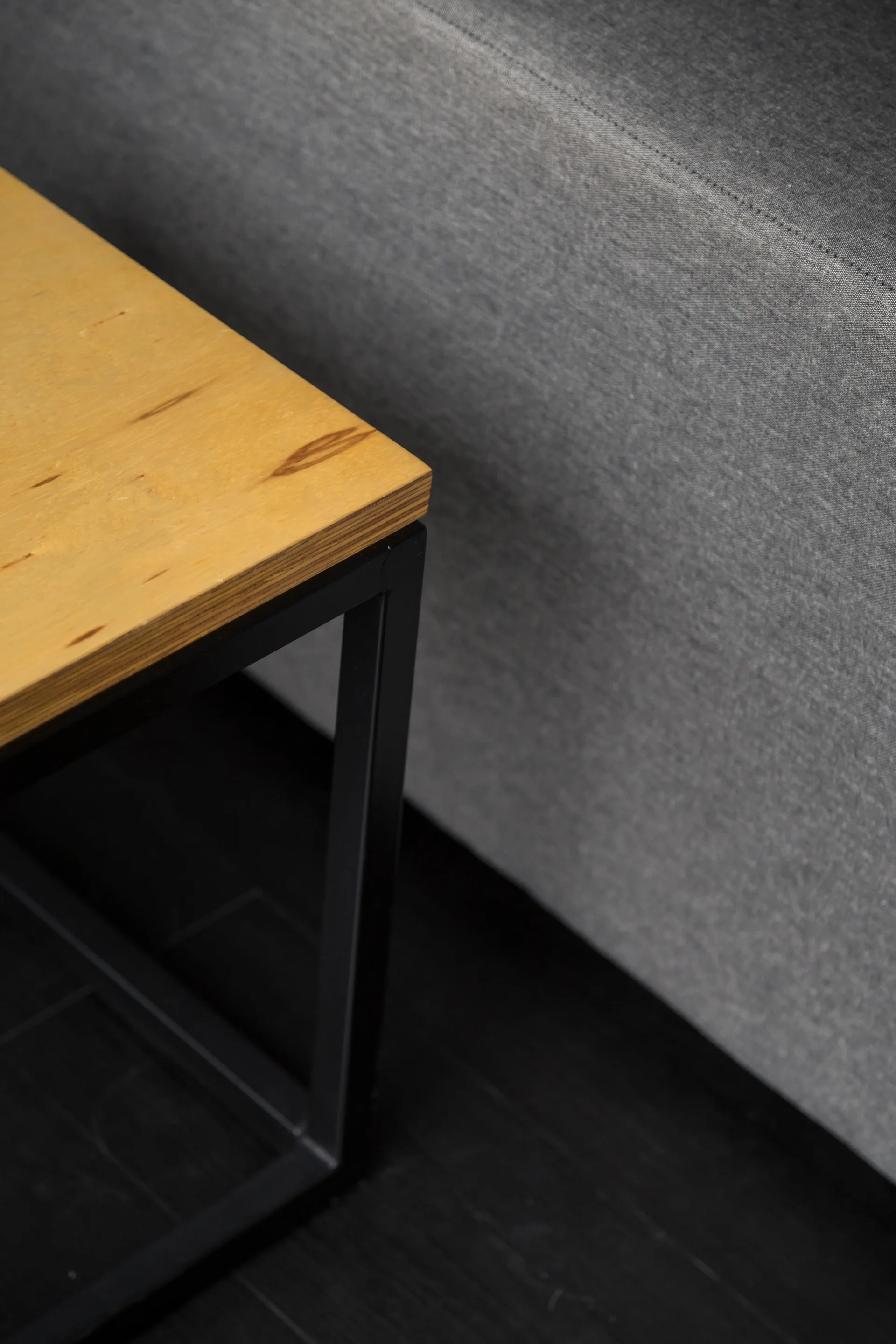 A close-up of a light wood tabletop with a black metal frame next to a gray fabric sofa on a dark wood floor.