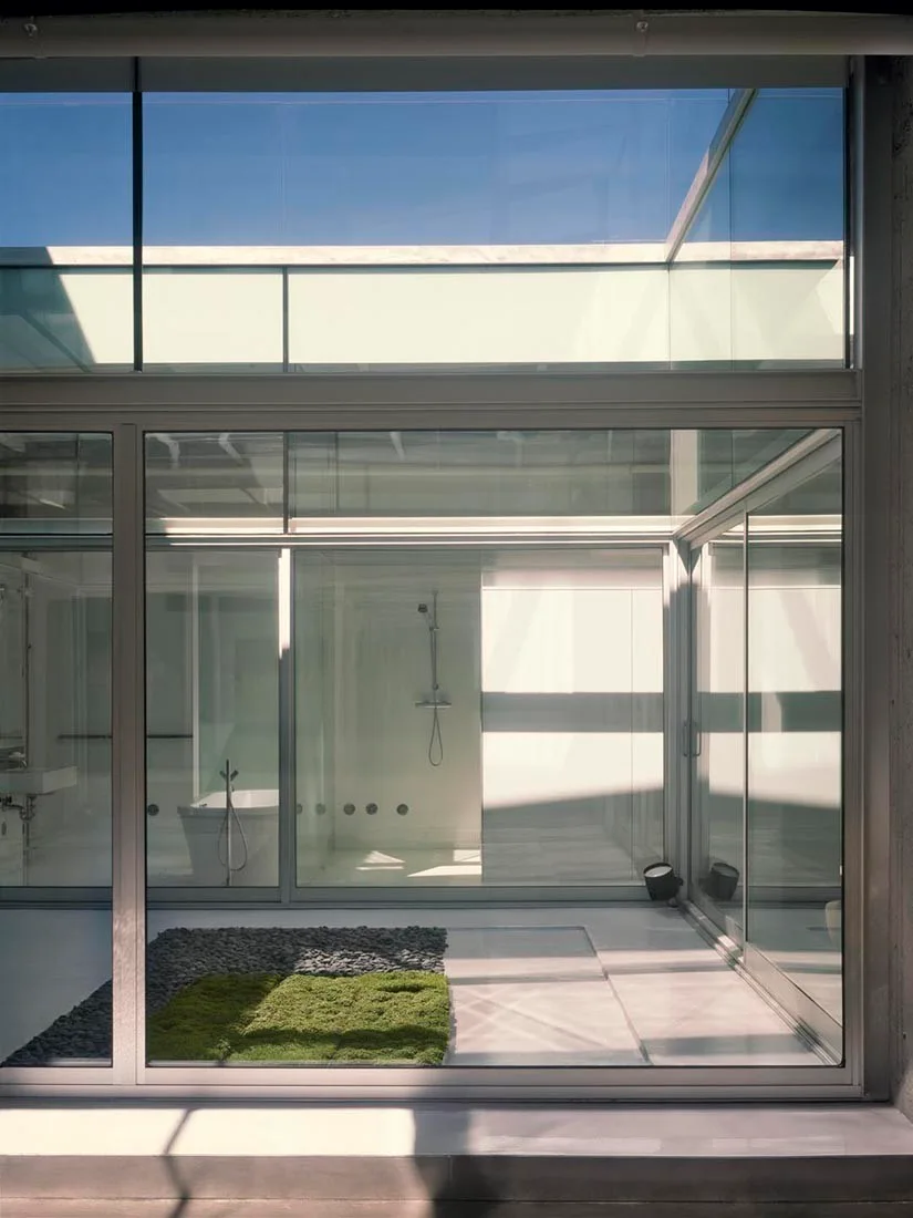 A modern white bathroom with a tub and shower is separated by large glass walls from an inner courtyard featuring green moss and grey stones.