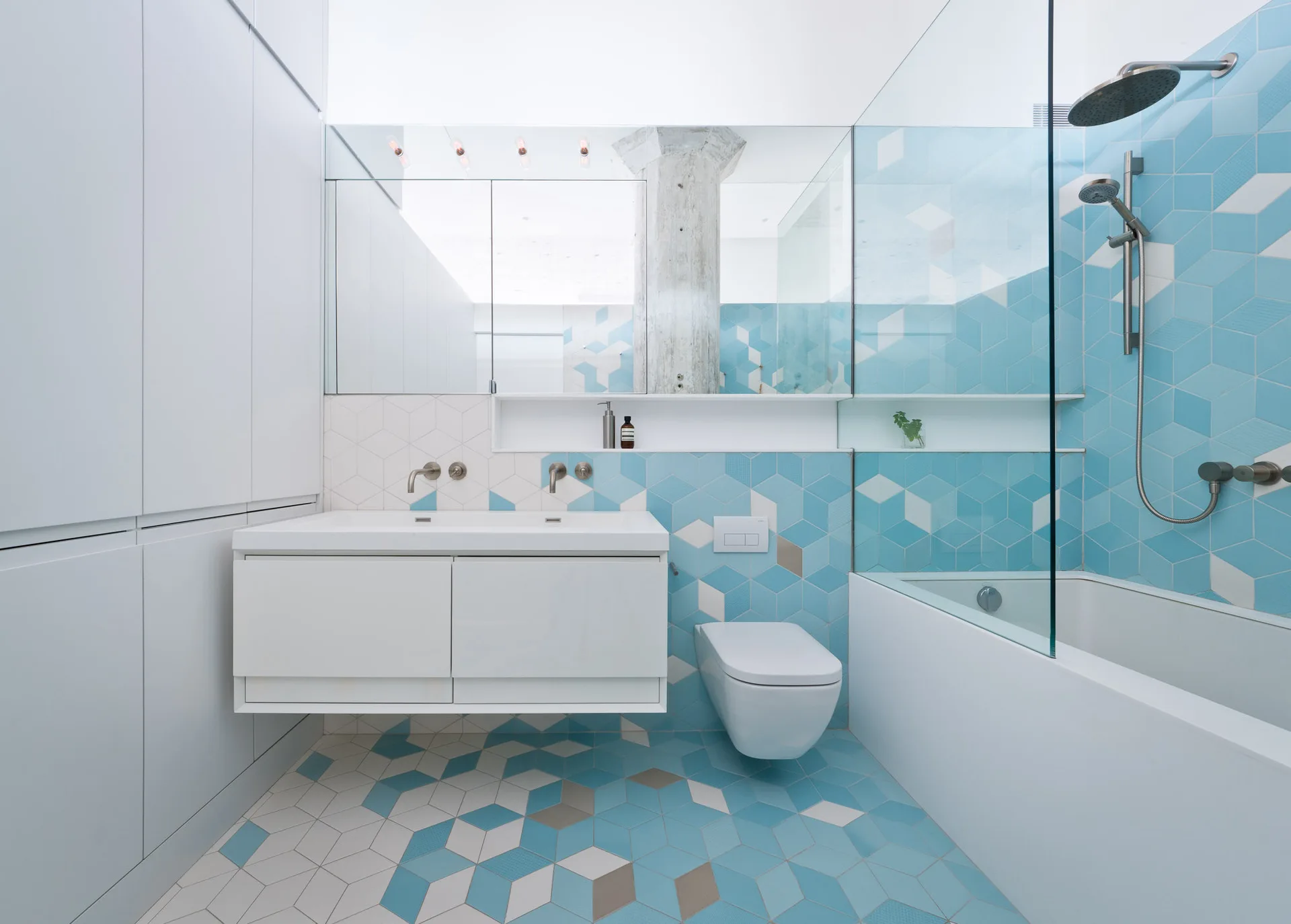 A modern bathroom features blue and white hexagonal tiles on the floor and walls, a white wall-mounted vanity, and a glass-enclosed shower.