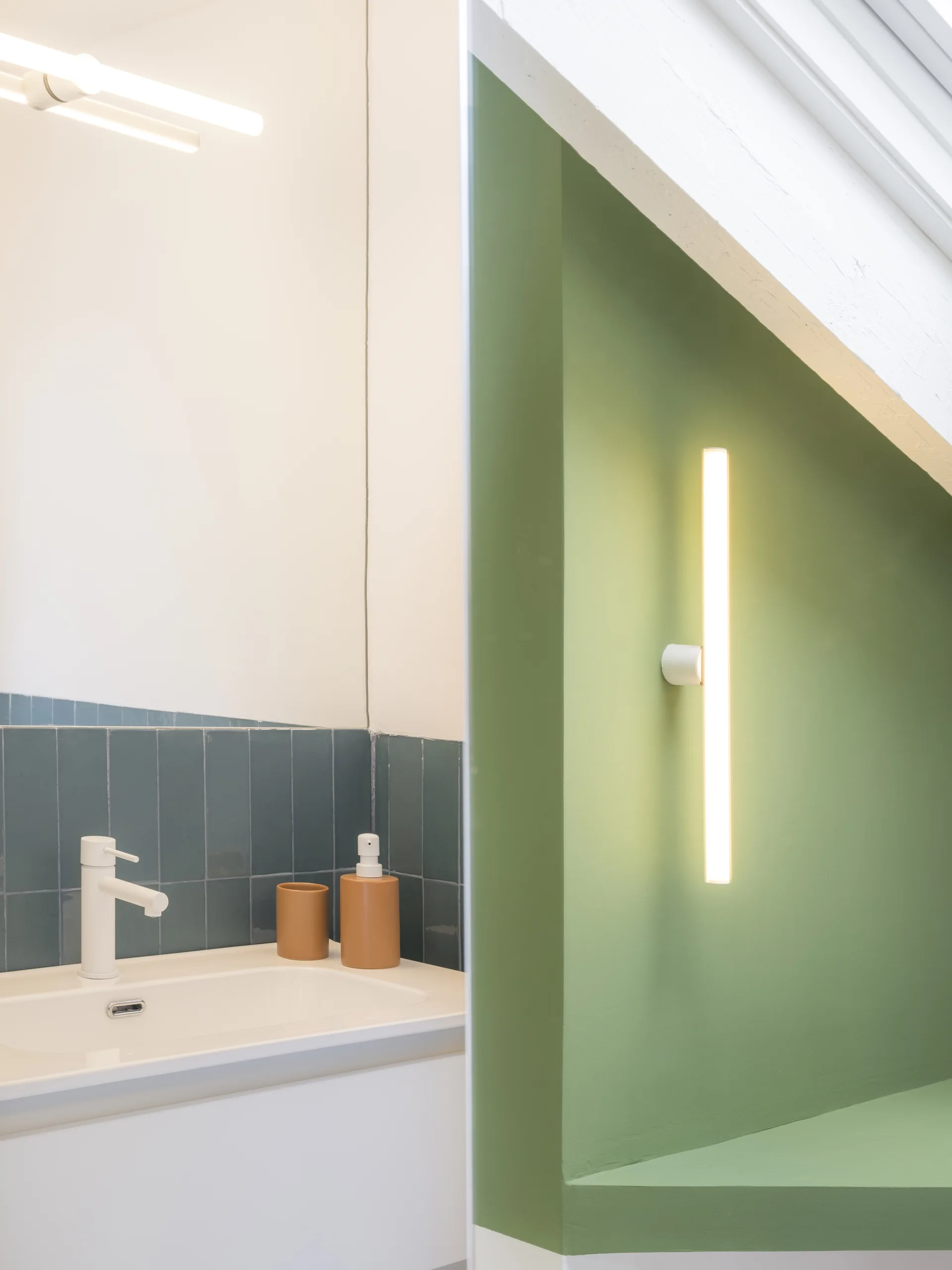 A modern minimalist bathroom vanity area with slate blue vertical tile, a white integrated sink and faucet, and a green wall with linear tube sconces.