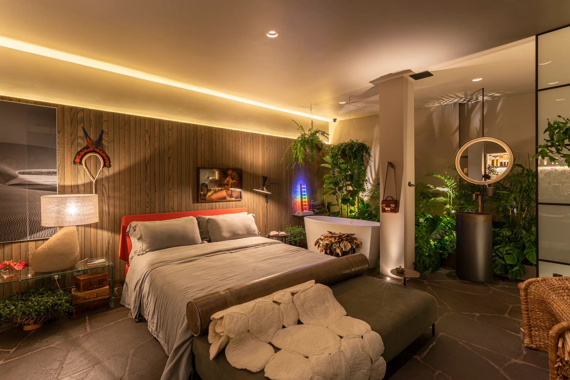 A contemporary bedroom suite features a bed against a wood-paneled wall, flagstone flooring, a freestanding bathtub, and lush green houseplants.