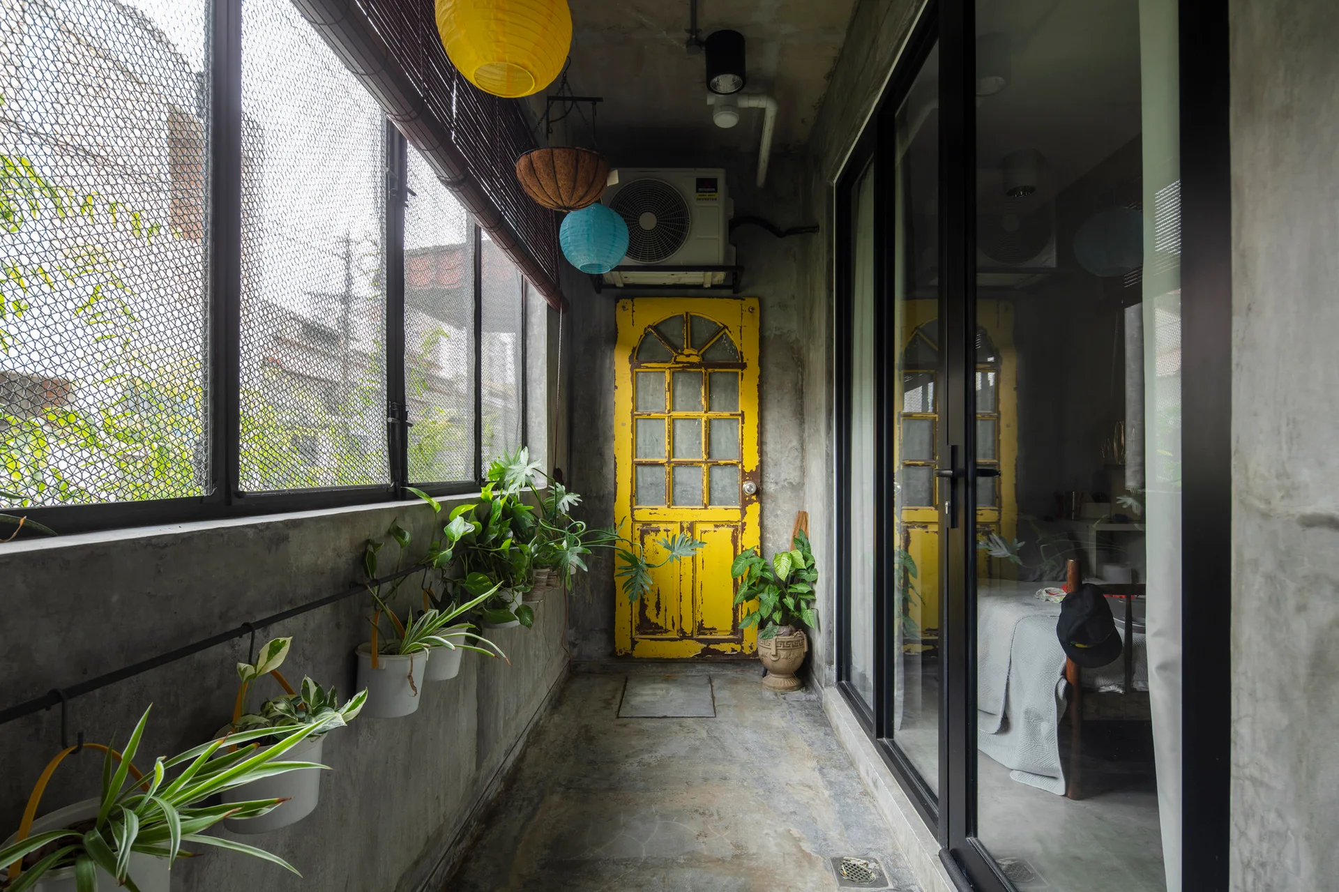 A narrow concrete balcony features a weathered yellow door, wall-mounted green plants, black-framed glass sliding doors, and hanging paper lanterns.