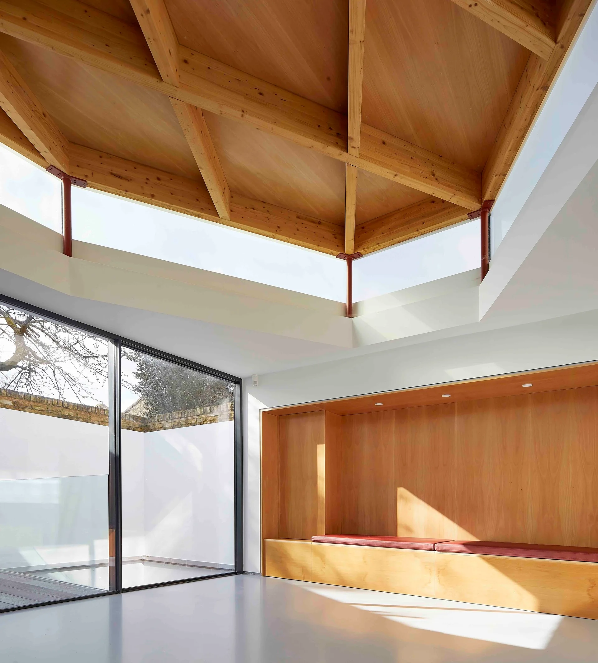 A bright minimalist room with a high vaulted timber ceiling, built-in wooden bench with a red cushion, and large glass sliding doors.