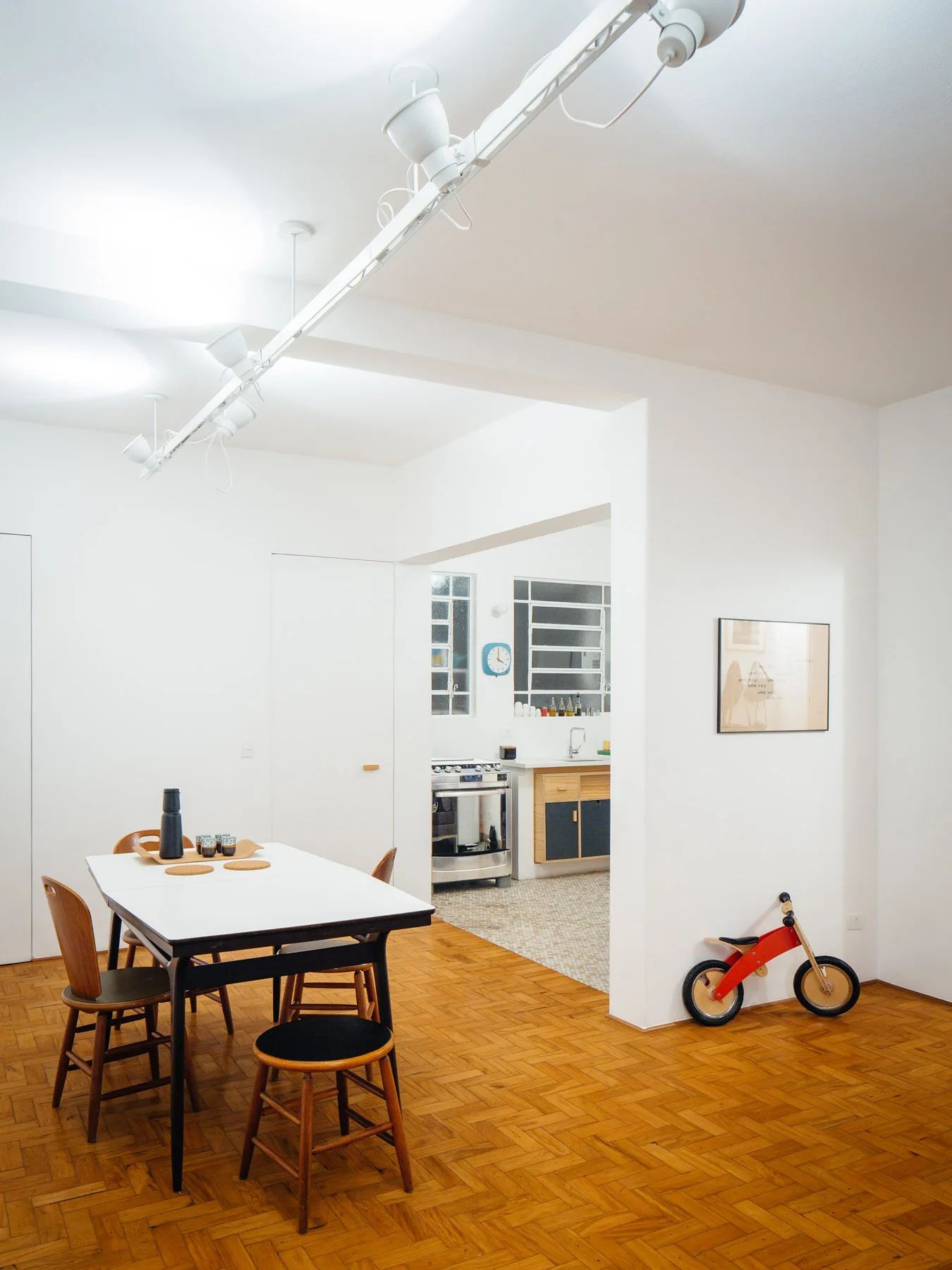 A minimalist dining area with a white table and wooden chairs features herringbone parquet floors and an opening into an adjacent kitchen.