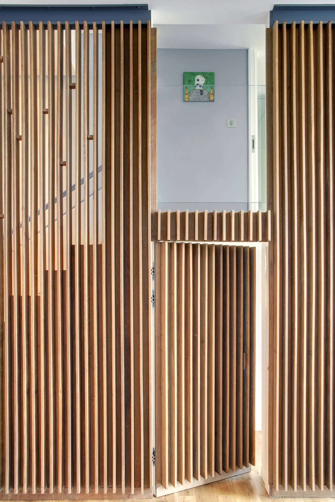 A vertical wooden slatted screen features an integrated hinged door that partially reveals a staircase and a light blue wall.