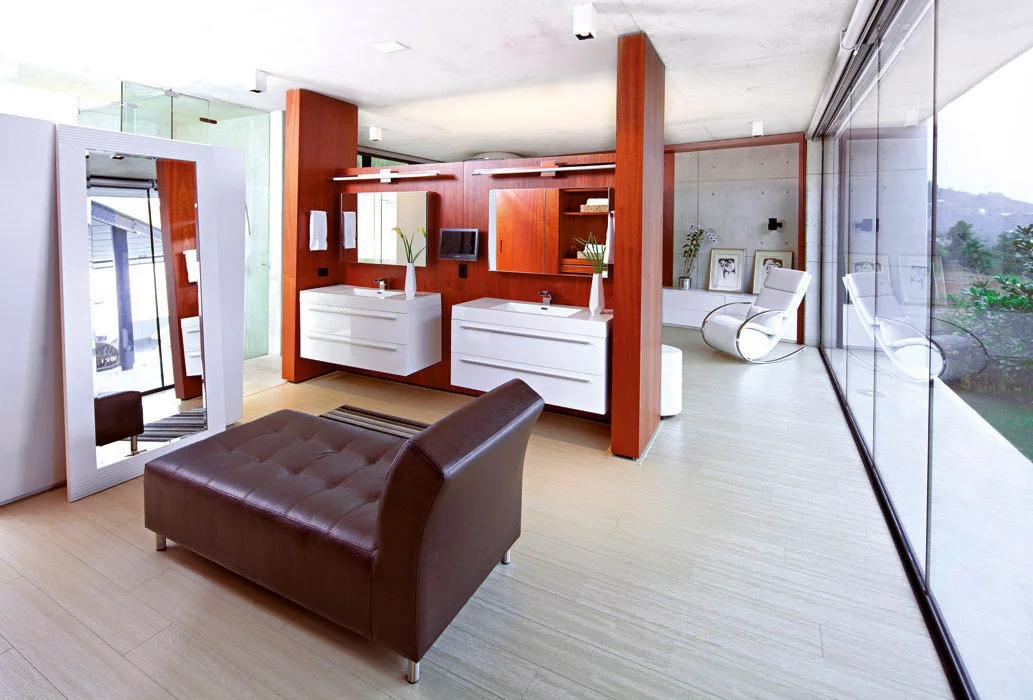 A spacious bathroom features a central wood divider wall holding floating white vanities, a brown leather chaise, and a floor-to-ceiling glass wall.