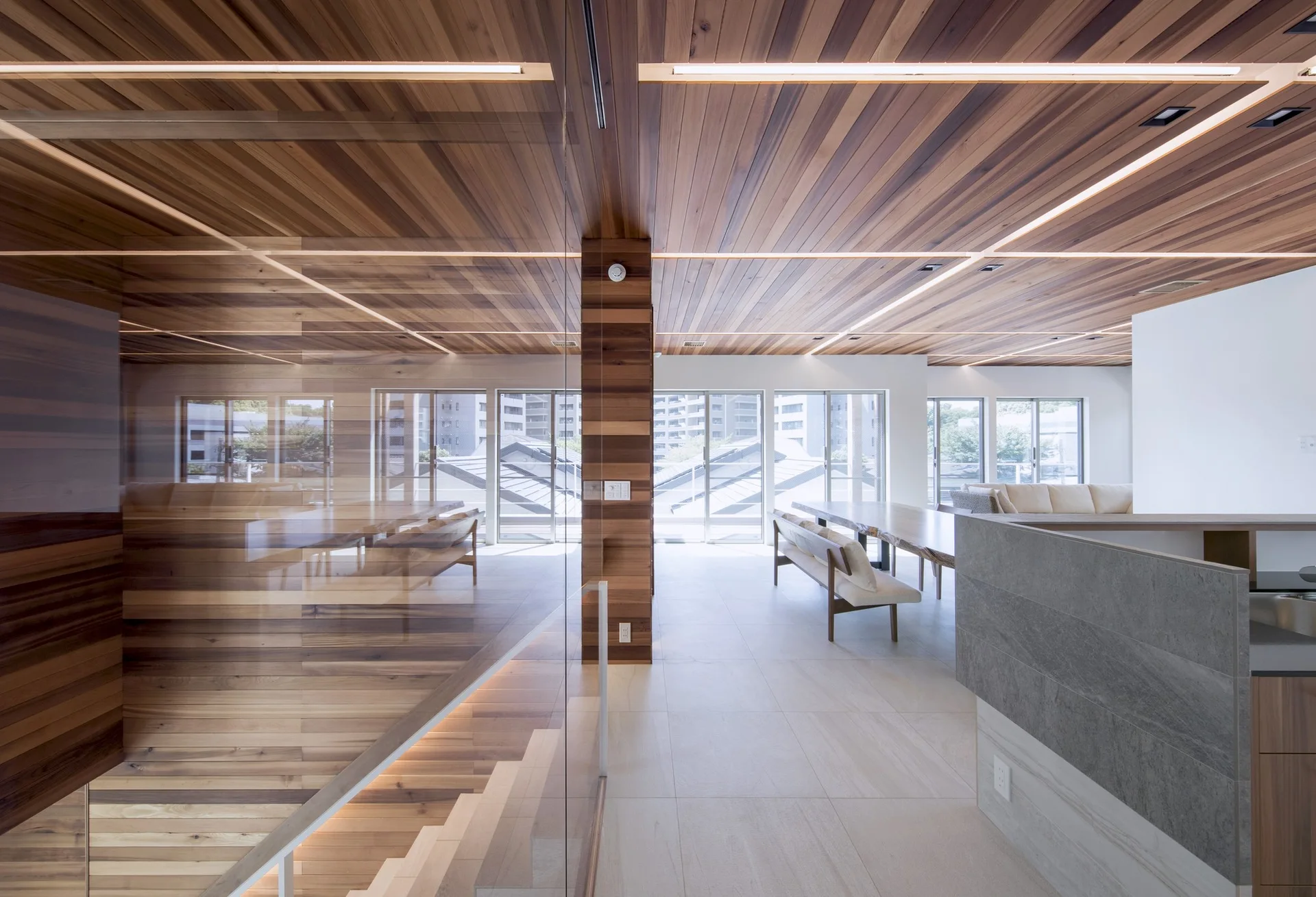A modern open-concept dining area features a slatted wood ceiling, a live-edge wooden table, large format floor tiles, and a glass partition.