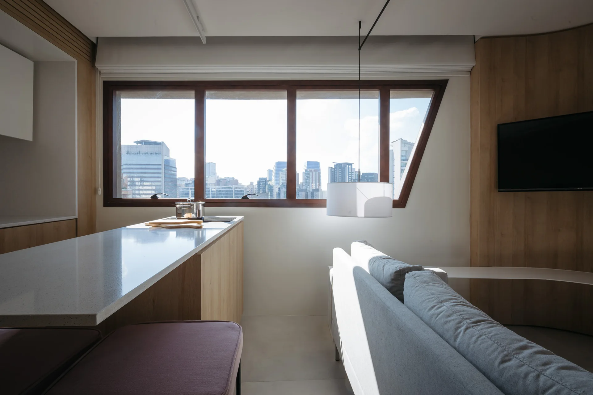 A modern living area featuring a grey sofa, a light wood kitchen island, and a large asymmetric window overlooking a city skyline.