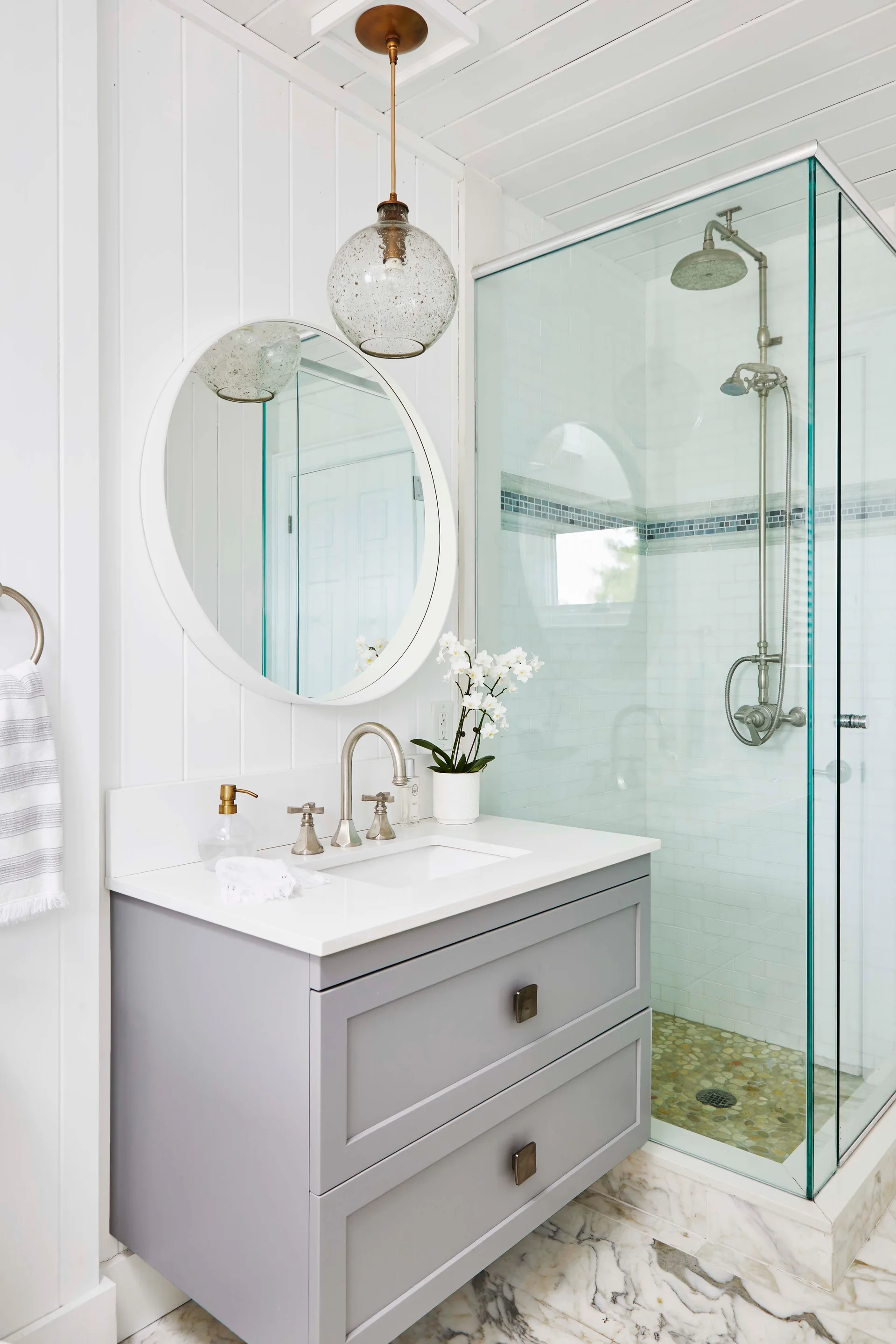A bright bathroom features a grey two-drawer vanity, a round mirror, a glass globe pendant light, and a glass-enclosed walk-in shower.