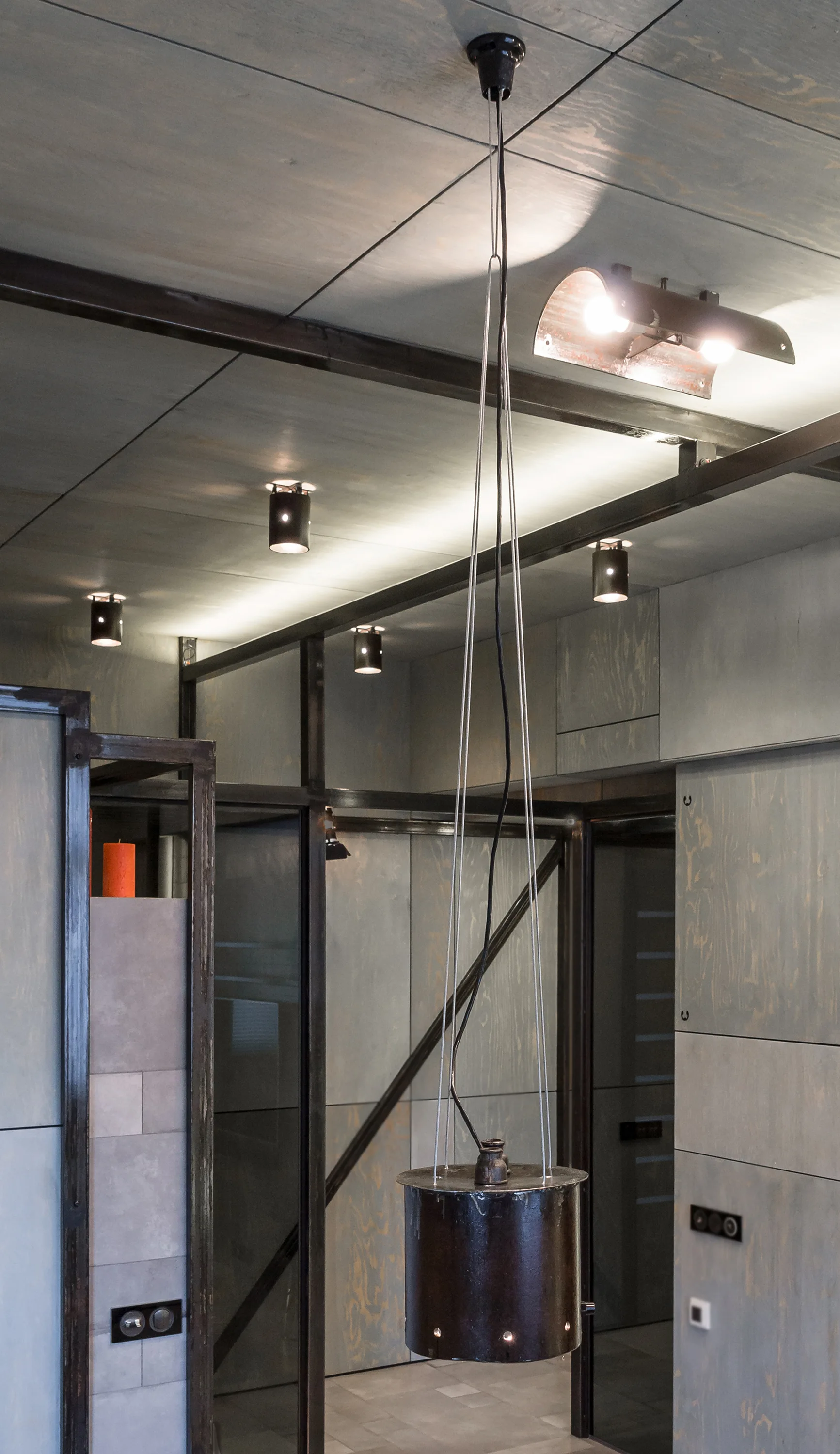 Grey stained plywood panels cover the walls and ceiling of an industrial room featuring dark steel structural beams and a hanging cylinder light.