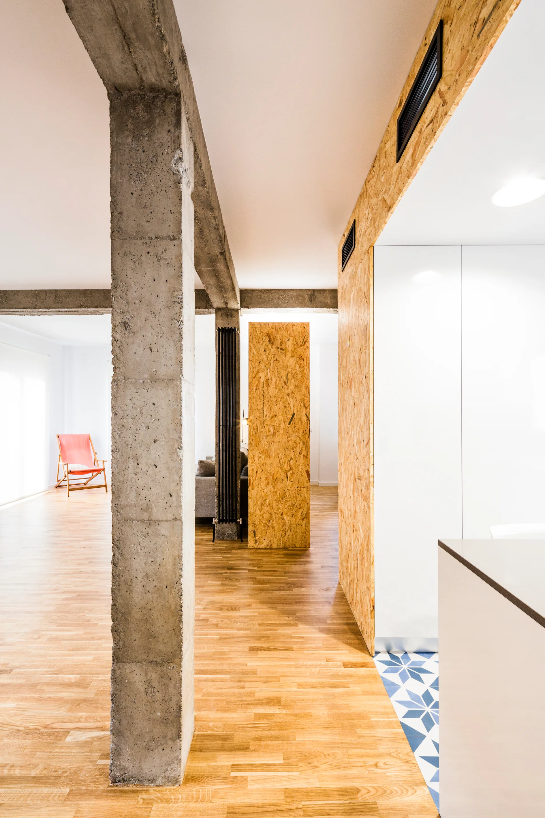 An open apartment interior showing a rough concrete pillar, light wood flooring, an OSB clad wall, and blue patterned tile floor inserts.