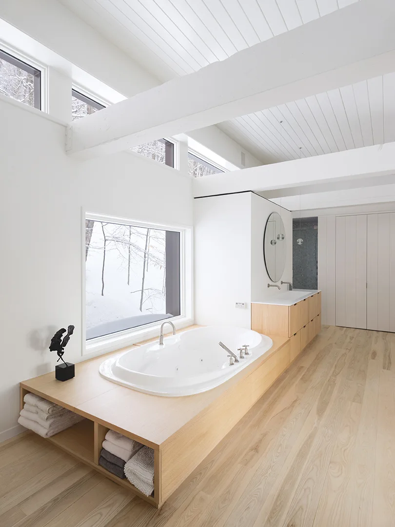 Minimalist bathroom featuring a light wood integrated bathtub and vanity, white vaulted ceiling with beams, and a window with a snowy view.