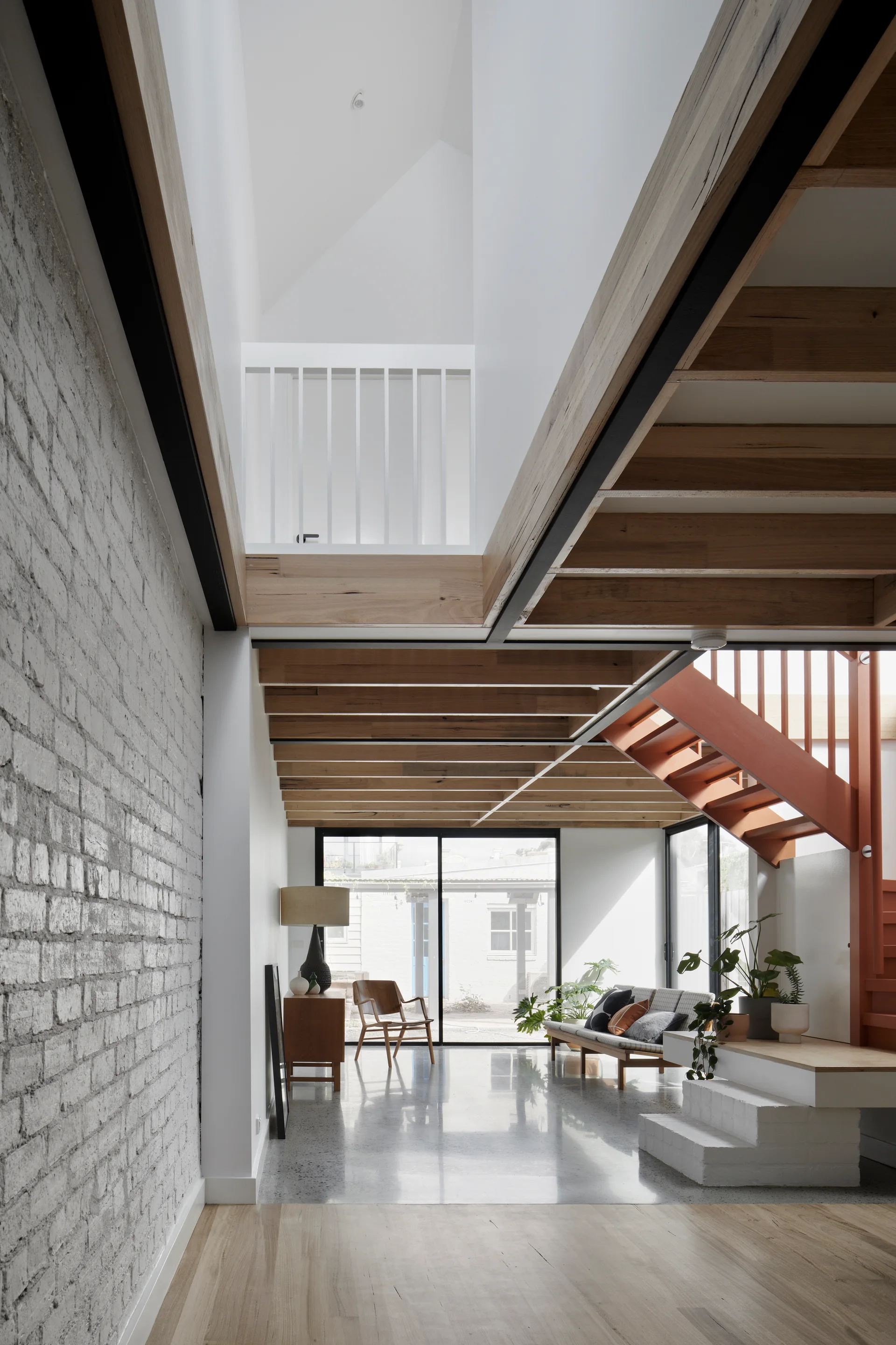A bright modern living room with exposed timber ceiling joists, a white painted brick wall, polished concrete floor, and an orange metal staircase.