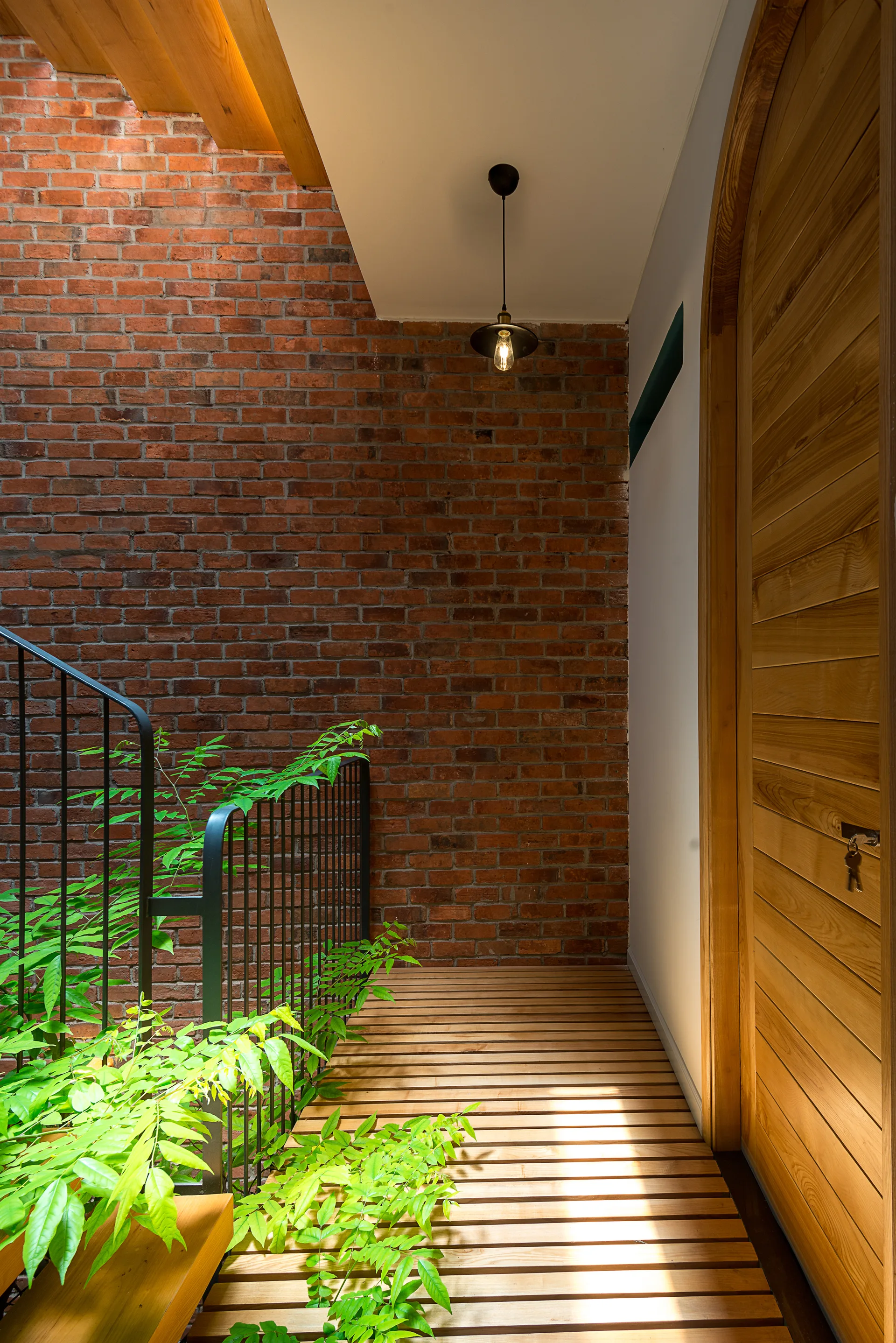 A staircase landing features an exposed red brick wall, a slatted wooden floor, an arched wood door, and a black metal railing with green plants.